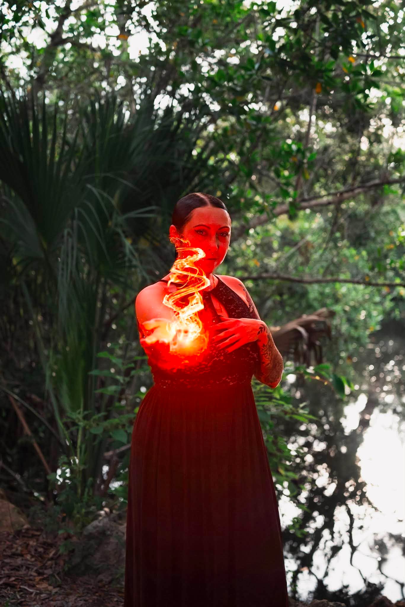 Woman standing outdoors near water, surrounded by trees and greenery, with sunlight filtering through leaves.