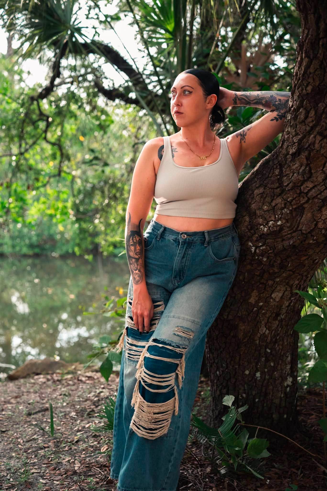 Woman with dark hair and tattoos leaning against a tree in a forested area, wearing a beige crop top and ripped jeans.