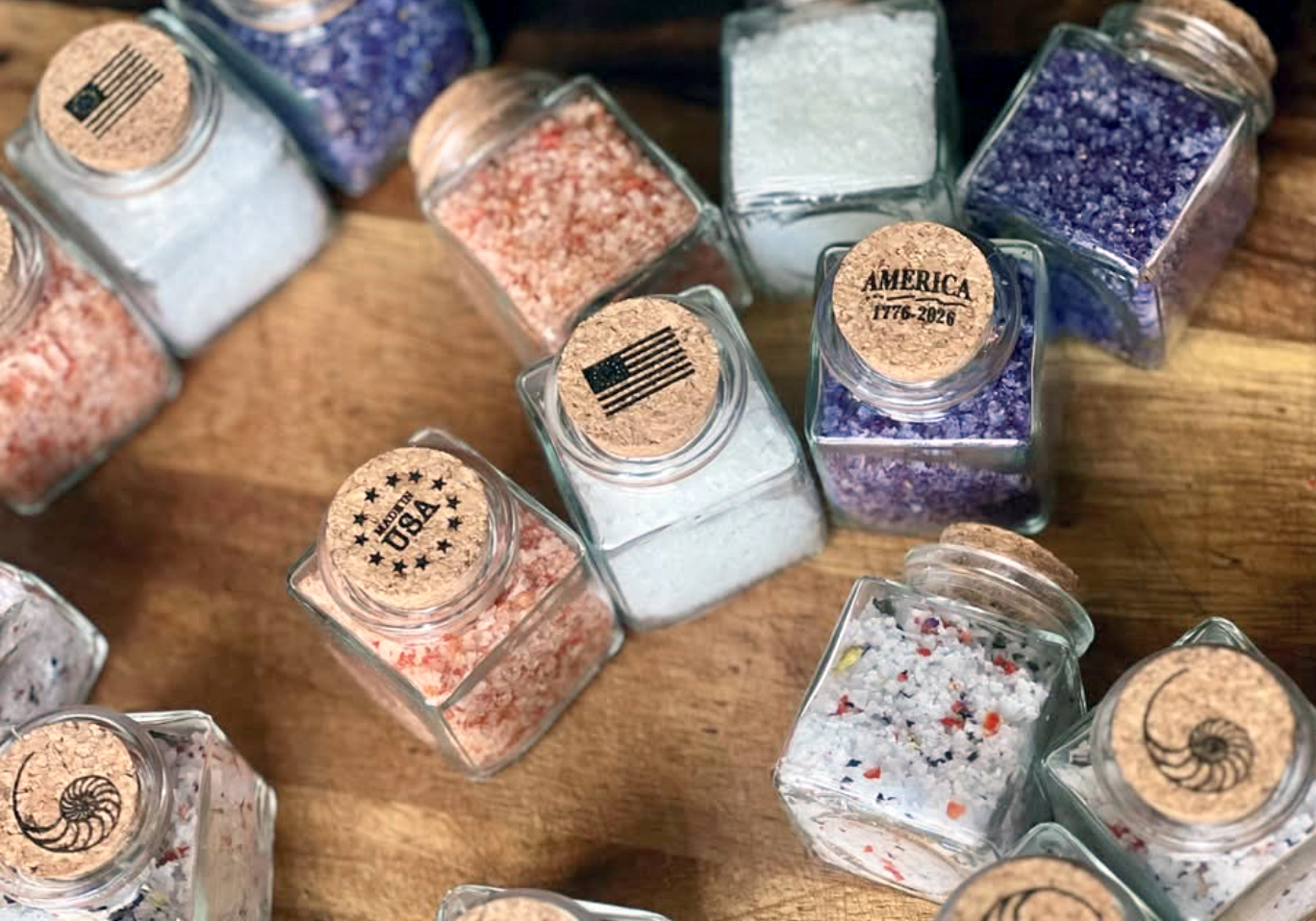 Small jars of colorful glitter on a wooden surface, some with labels and American flags, in various colors and textures.