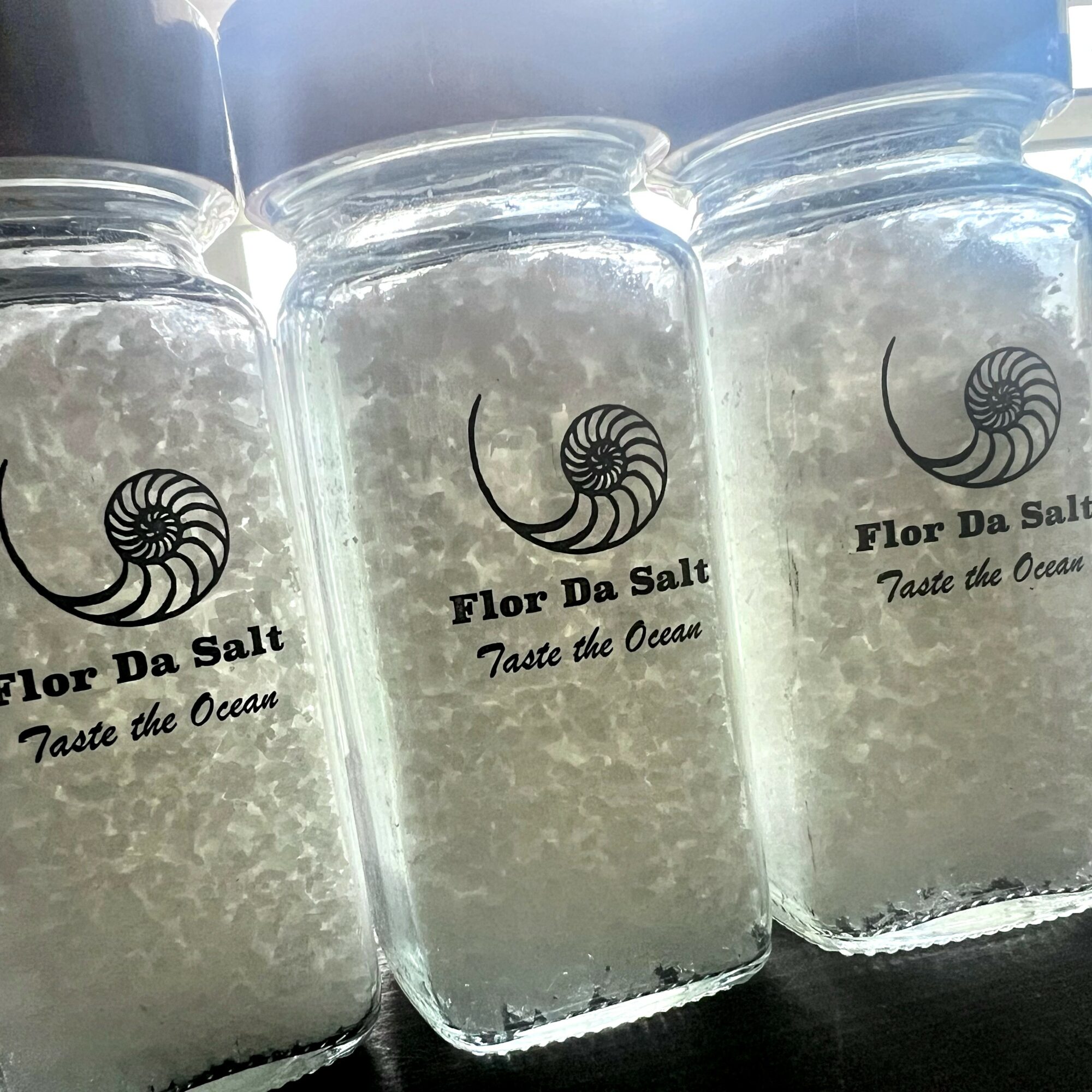 Three glass jars filled with salt, labeled 'Flor Da Salt' and 'Taste the Ocean,' arranged in a row.