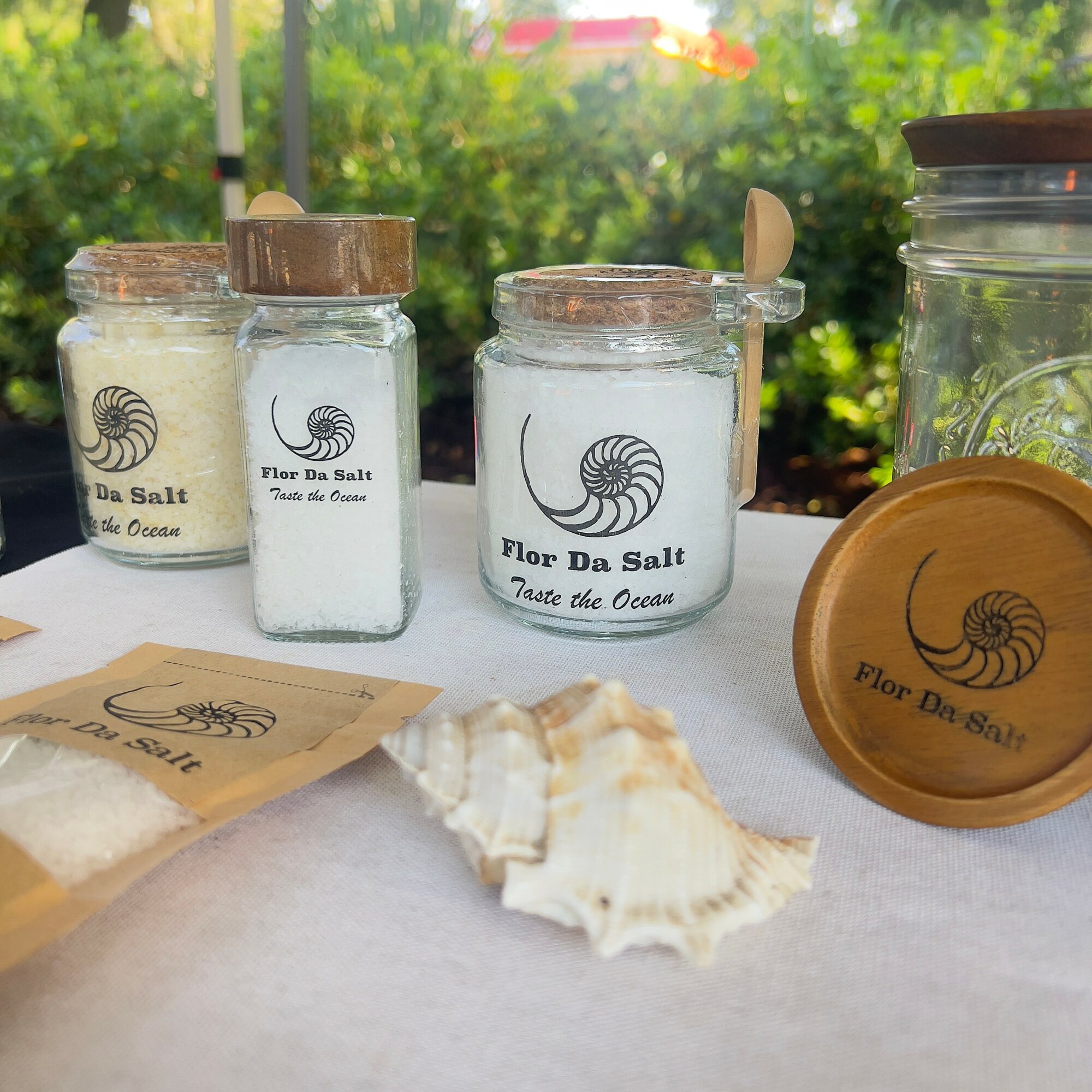 Three jars of salt with wooden lids, a shell, and a paper bag on a table outdoors with greenery in the background.