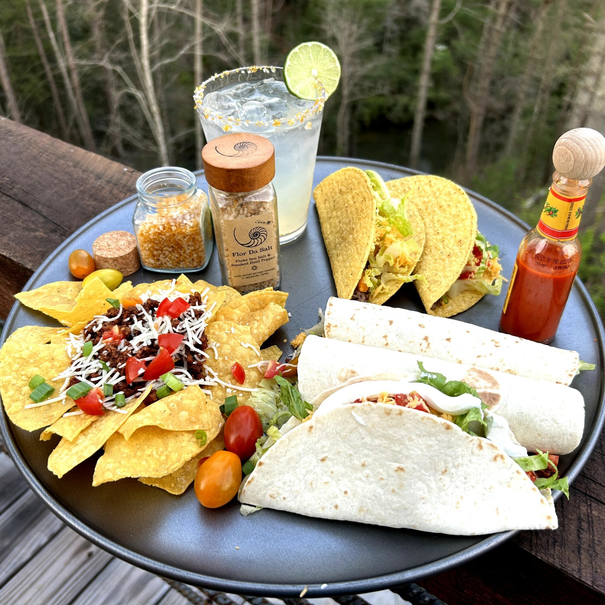 Plate with tacos, nachos, salad, and drinks outdoors, with trees in the background.