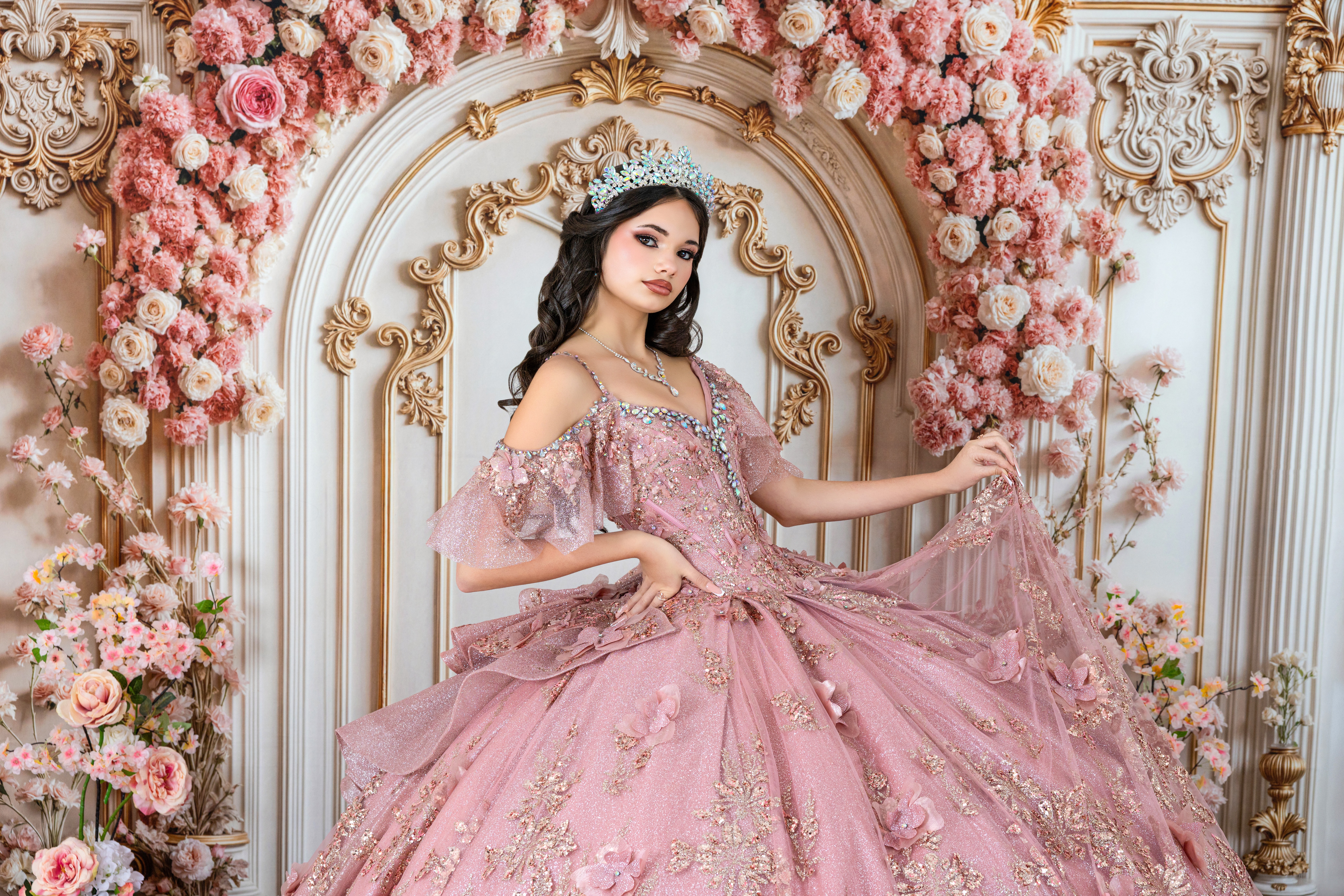Young woman in pink gown with floral background and ornate gold details, holding part of her dress, looking at camera.