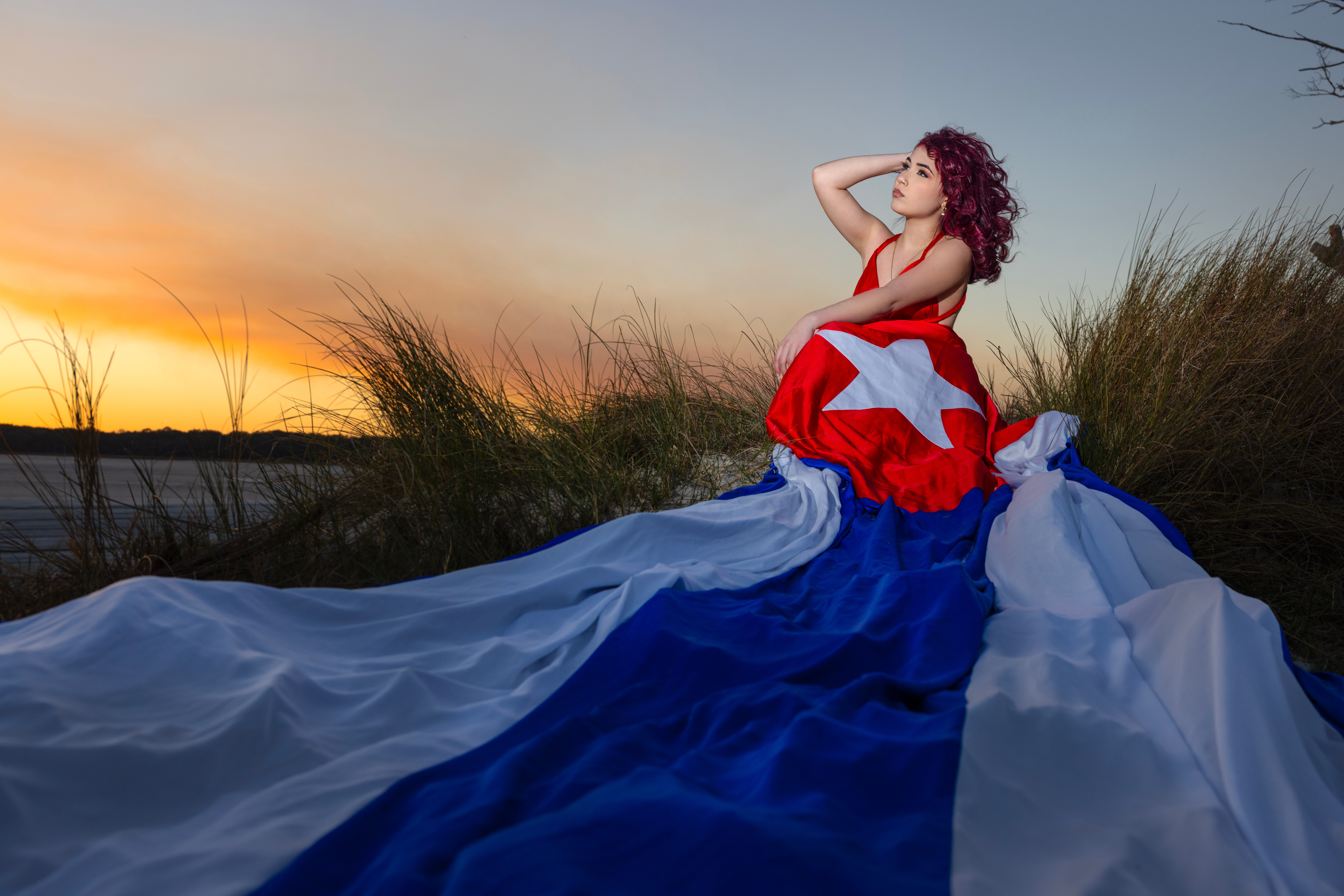 Woman with curly hair in a red, white, and blue dress sitting on grass at sunset, with a long flowing train.