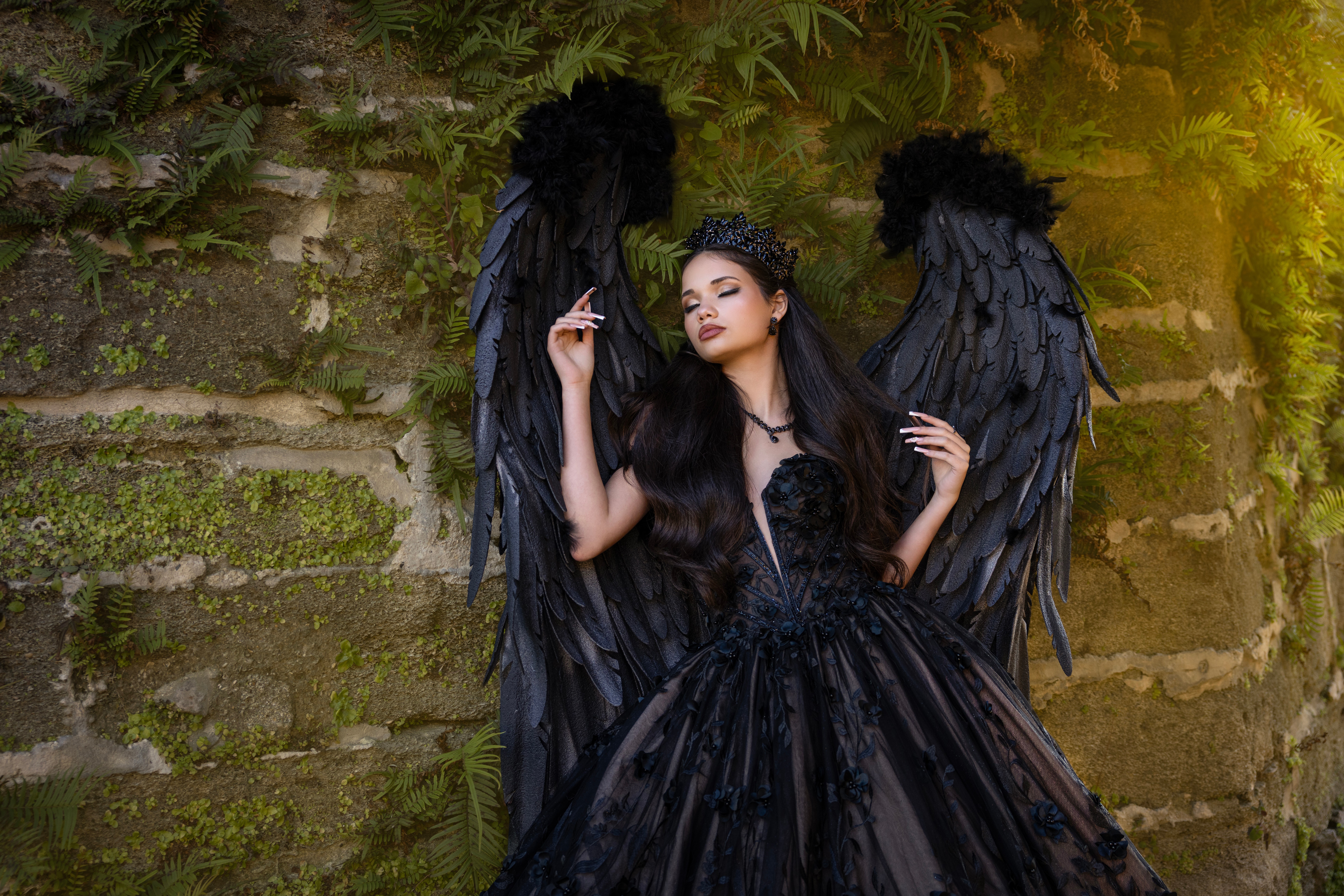 A woman with black wings and long dark hair stands against a mossy brick wall, wearing a black dress.