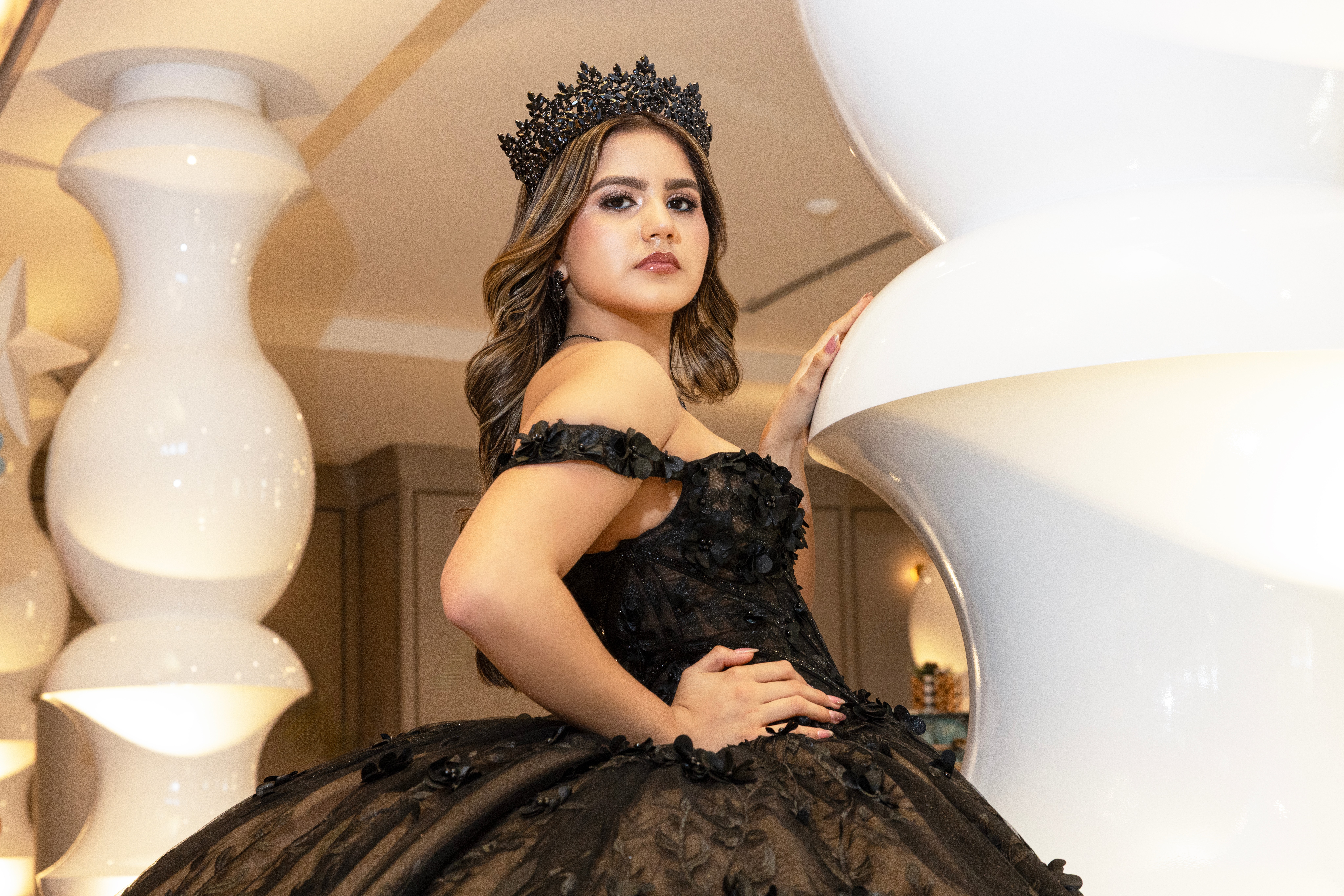 Young woman in a black dress and crown, posing indoors with large white decorative objects around her.