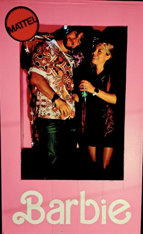 Two people talking, one with a patterned shirt and the other with a black dress, against a pink background with Barbie logo.