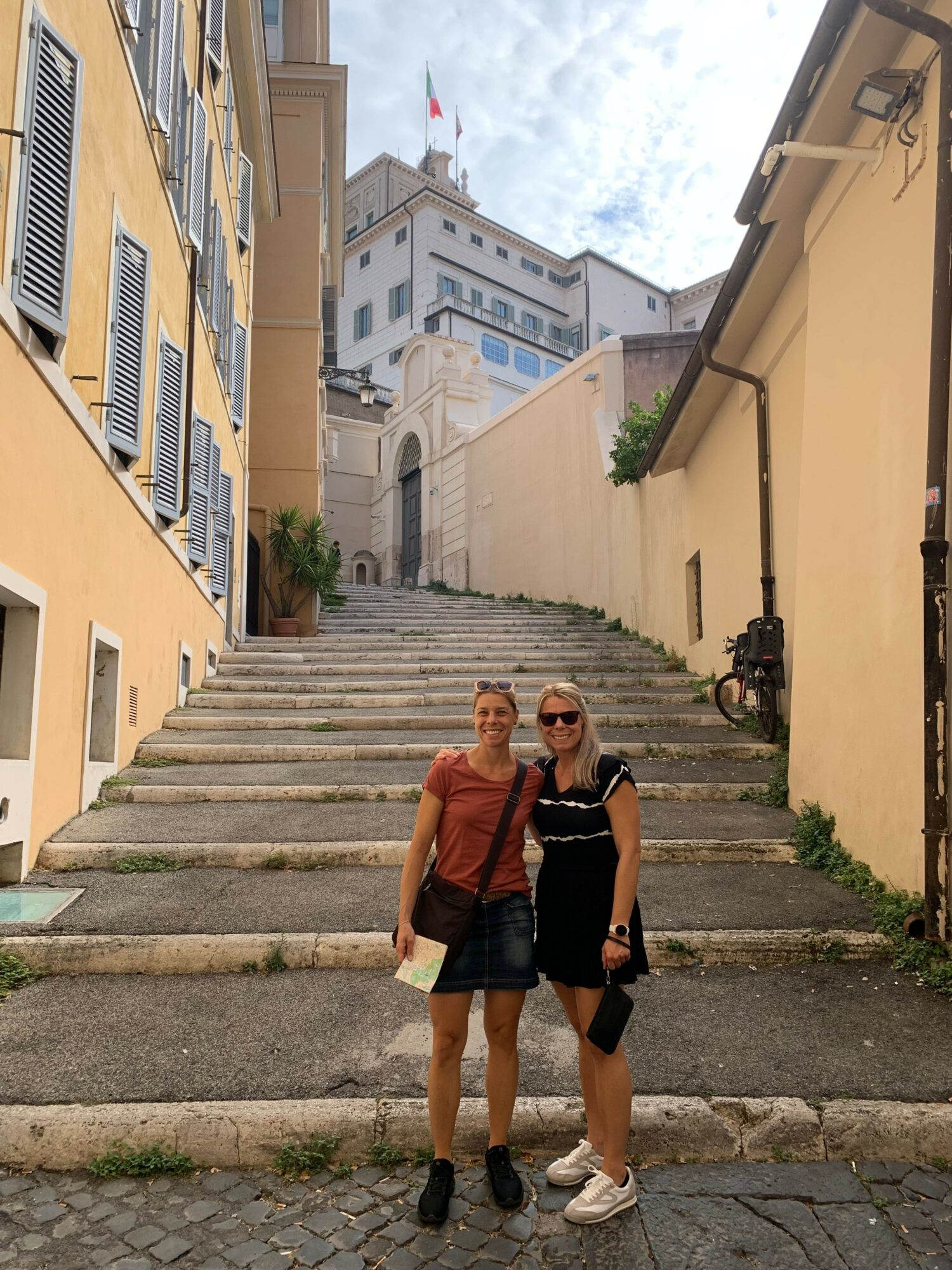 Two people walking on a stone staircase in an urban area, with buildings and a cloudy sky in the background.