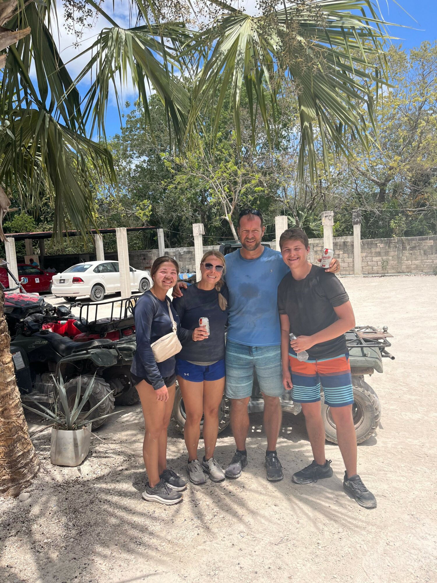 Four people stand together outdoors near palm trees and parked cars, smiling at the camera.