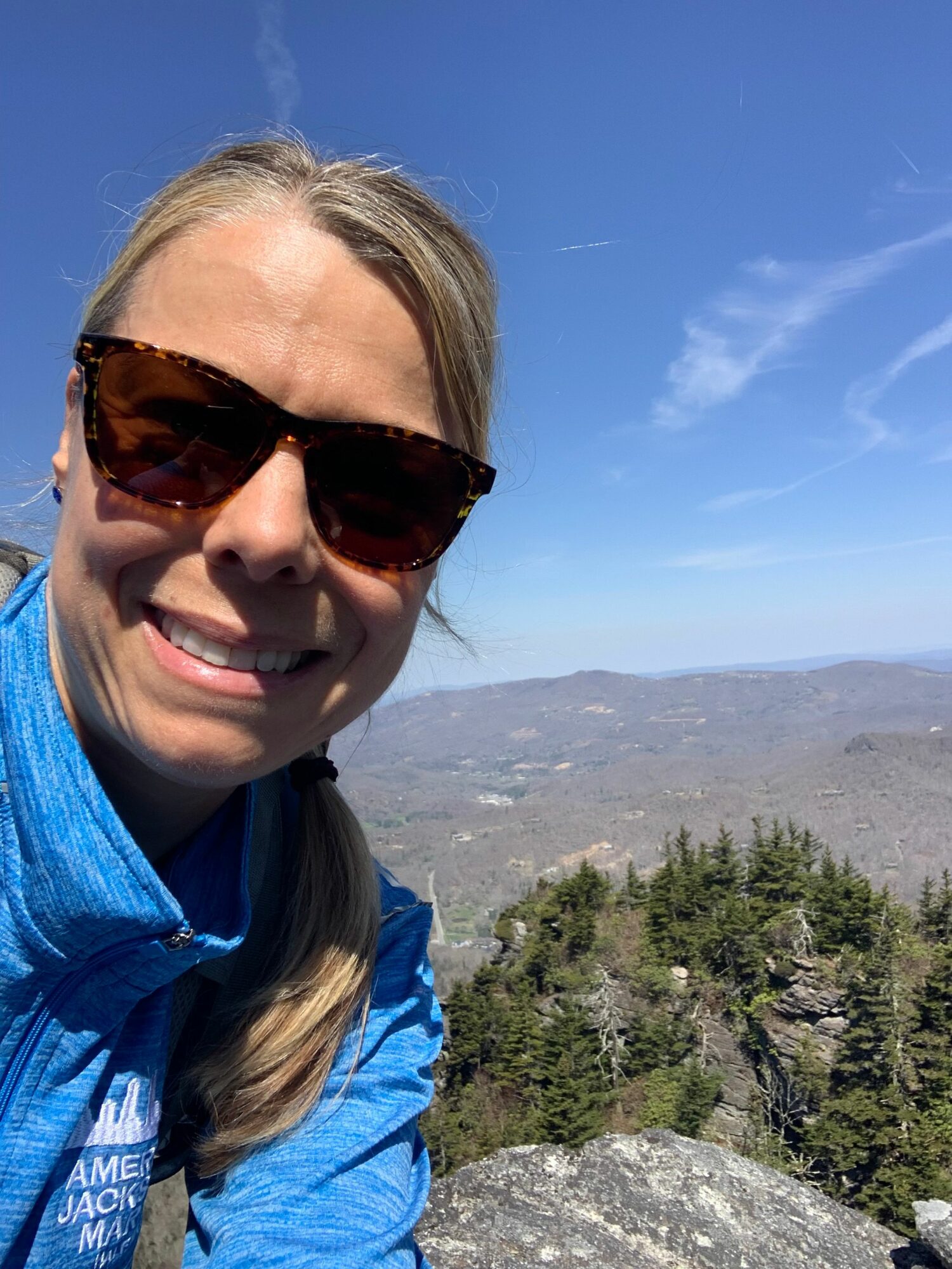 Woman wearing sunglasses and a blue jacket outdoors with a mountain and sky in the background.