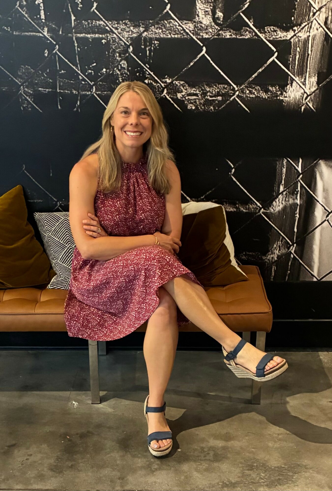 Woman with long blonde hair sitting on a brown sofa, smiling, wearing a pink patterned dress and sandals, in front of a black and white abstract wall.