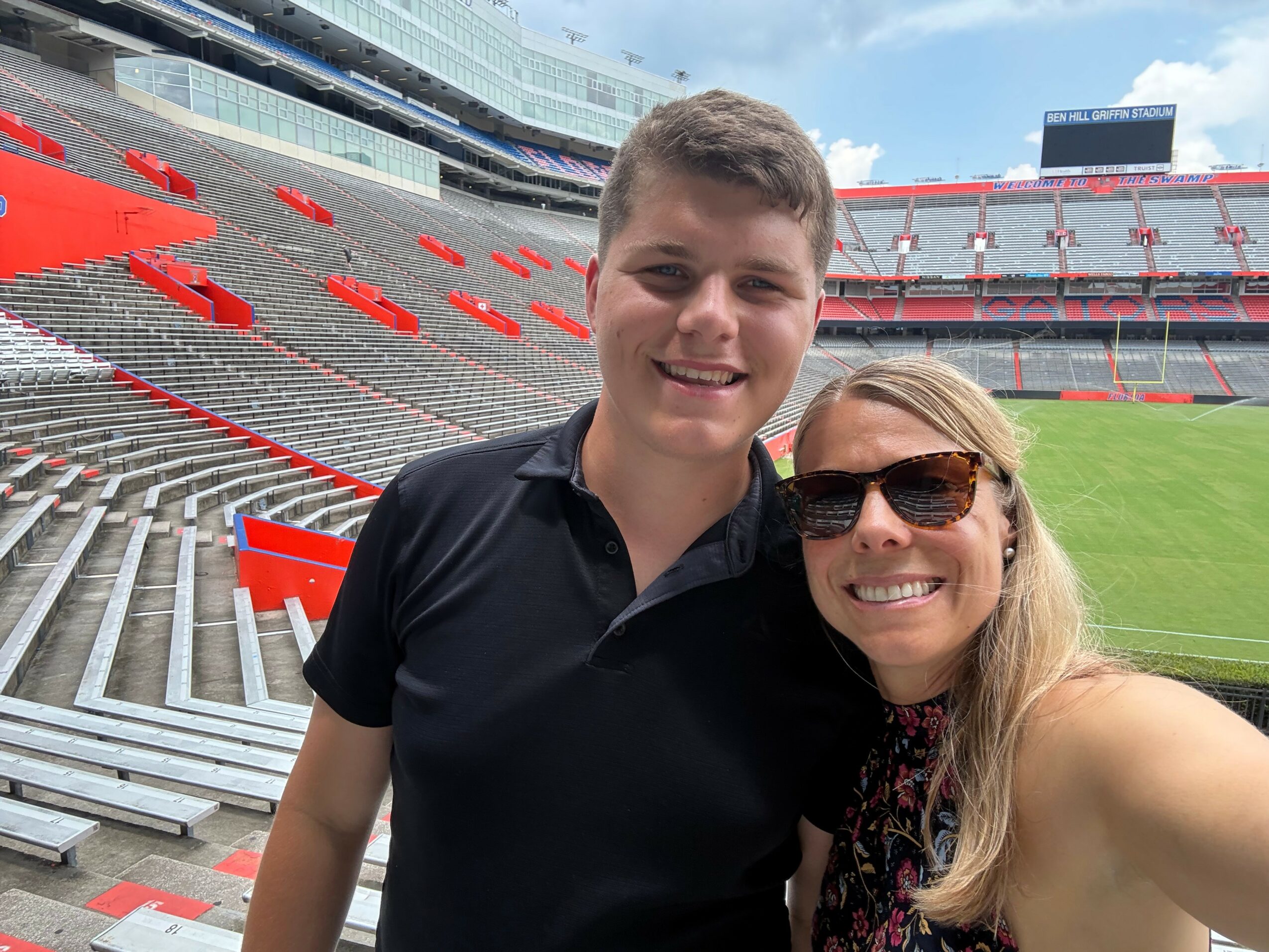 A young man and woman smiling at a stadium with empty seats and a field in the background.