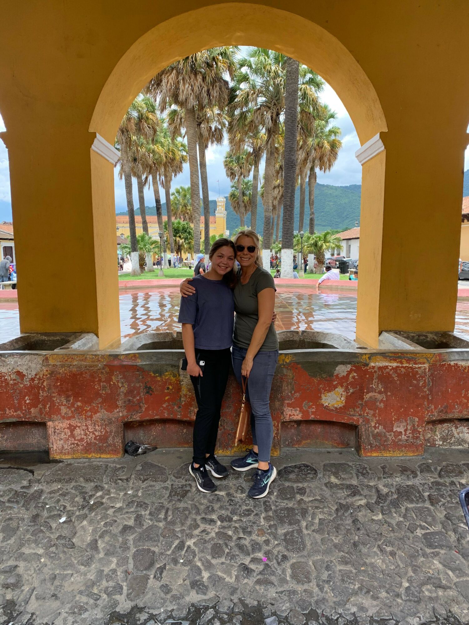 Two people standing under a yellow archway with trees and a body of water in the background.