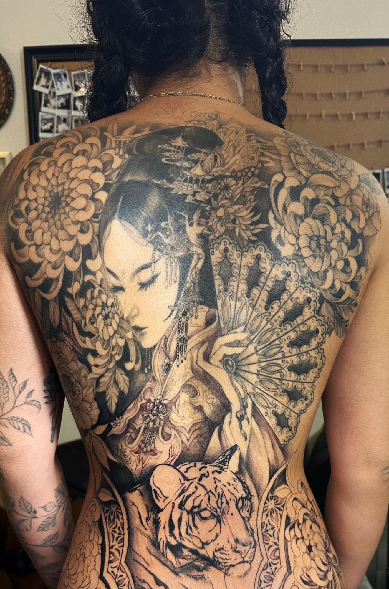 Back tattoo featuring a woman's face, floral patterns, a tiger's face, and a skull with a fan, in black and gray ink.