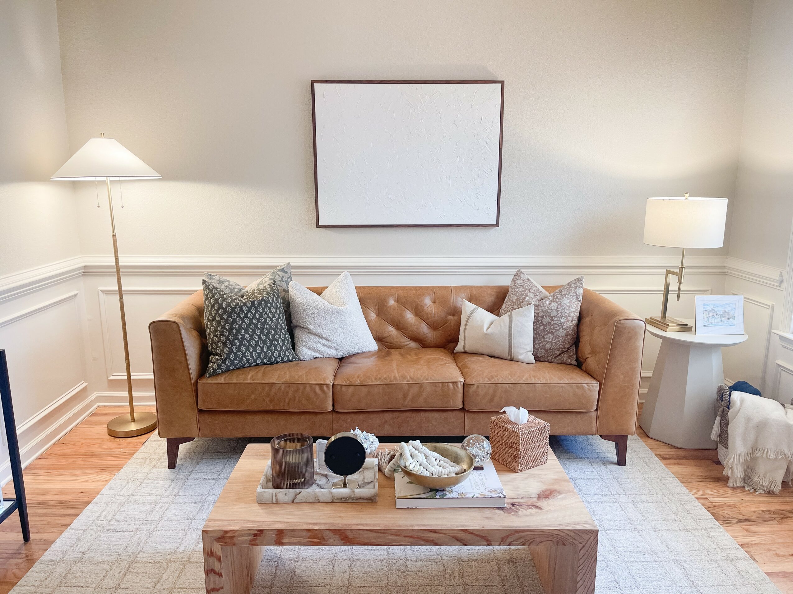 Living room with a brown sofa, two lamps, a coffee table, and a blank wall art, with a rug and hardwood floor.