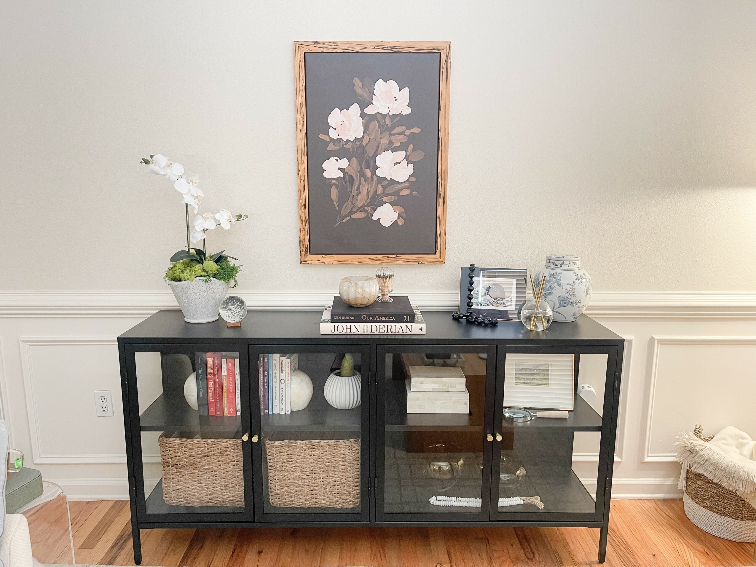 Black display cabinet with glass doors, decorative items, and books, against a light-colored wall with framed artwork above.