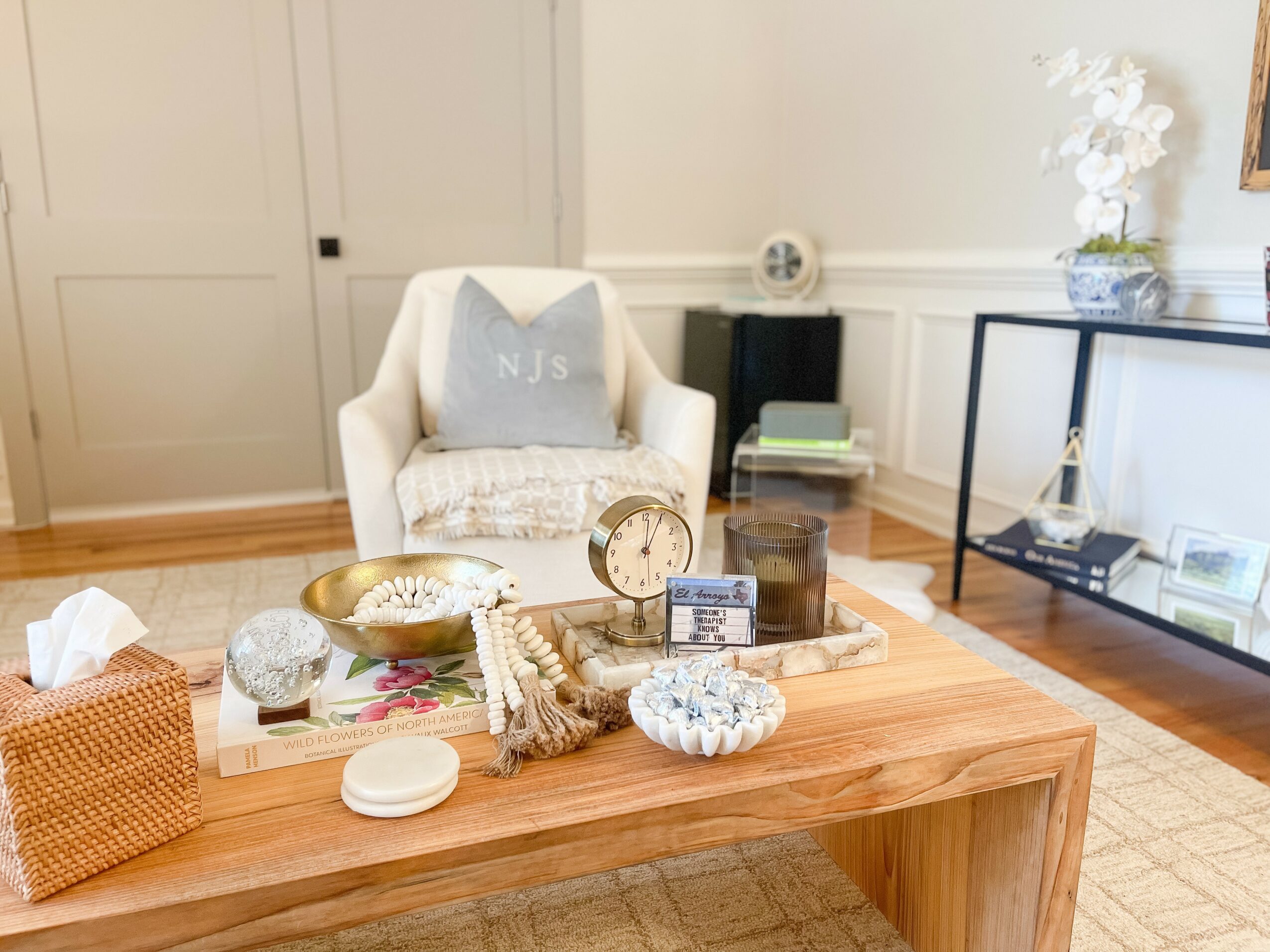 Living room with a white armchair, wooden coffee table, and a glass display shelf, decorated with flowers and books.