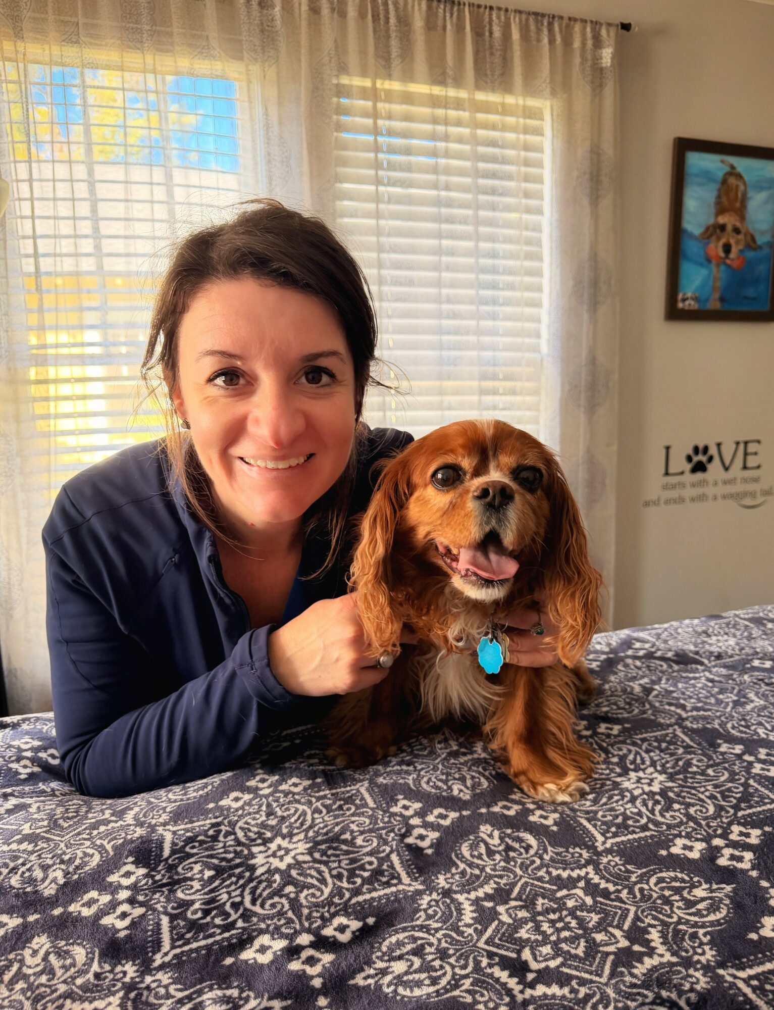 Woman with dark hair smiling, resting her chin on a brown dog with long ears on a patterned tablecloth.