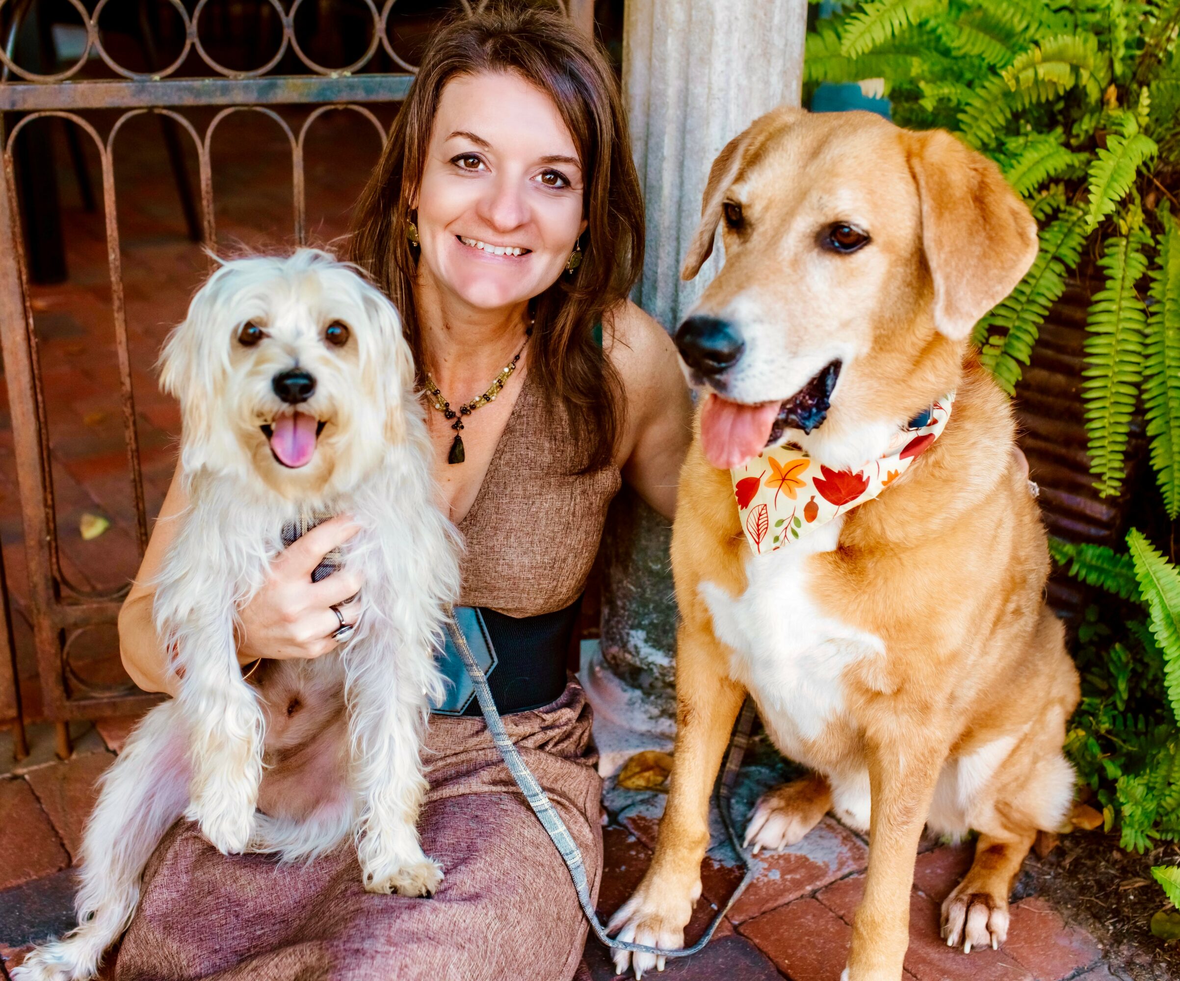 Woman smiling with two dogs, one small and one large, outdoors with greenery and a wooden fence behind.