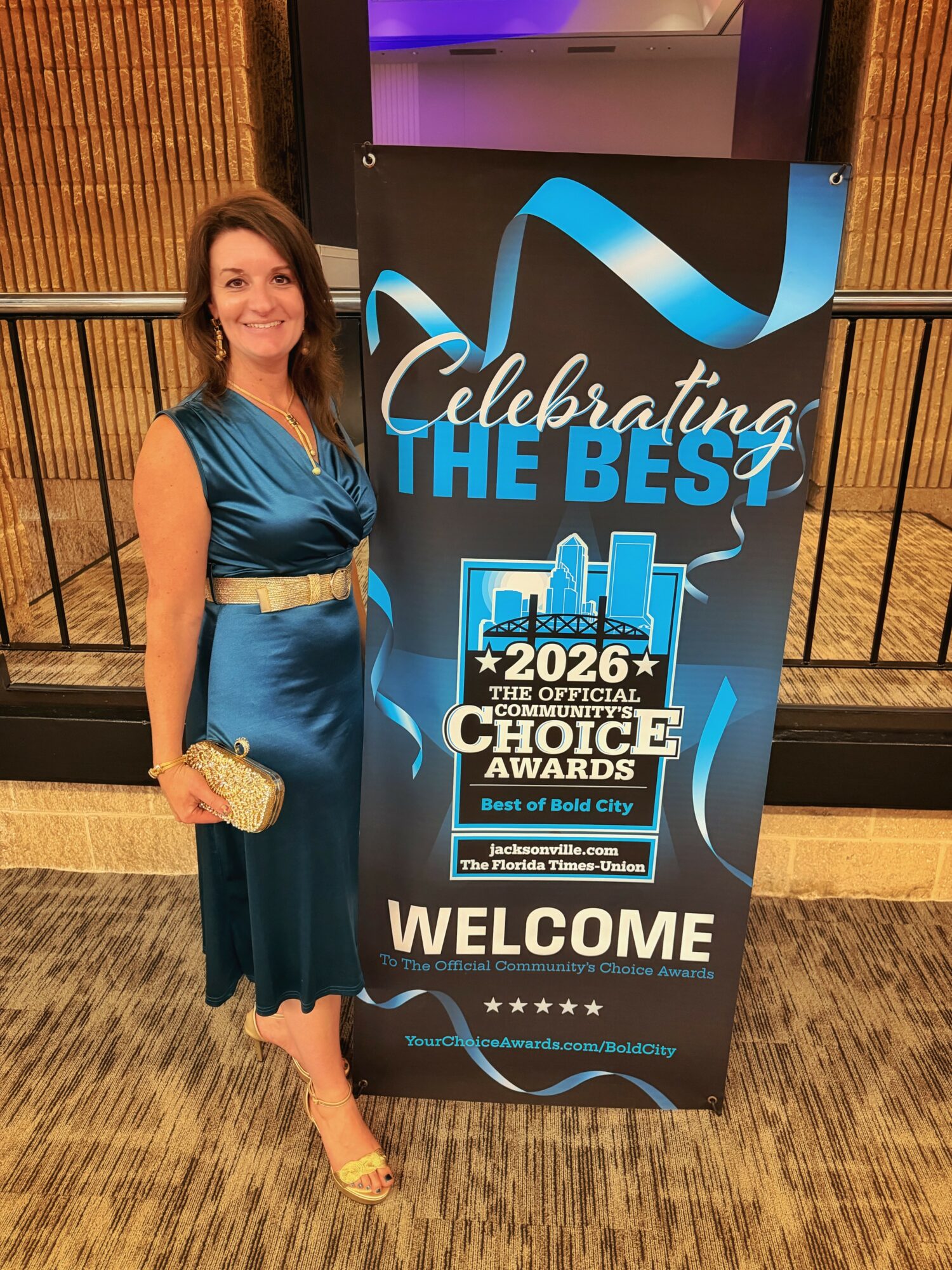 Woman in blue dress holding a clutch standing next to a sign for the 2026 Choice Awards, celebrating the best of bold city, indoors.