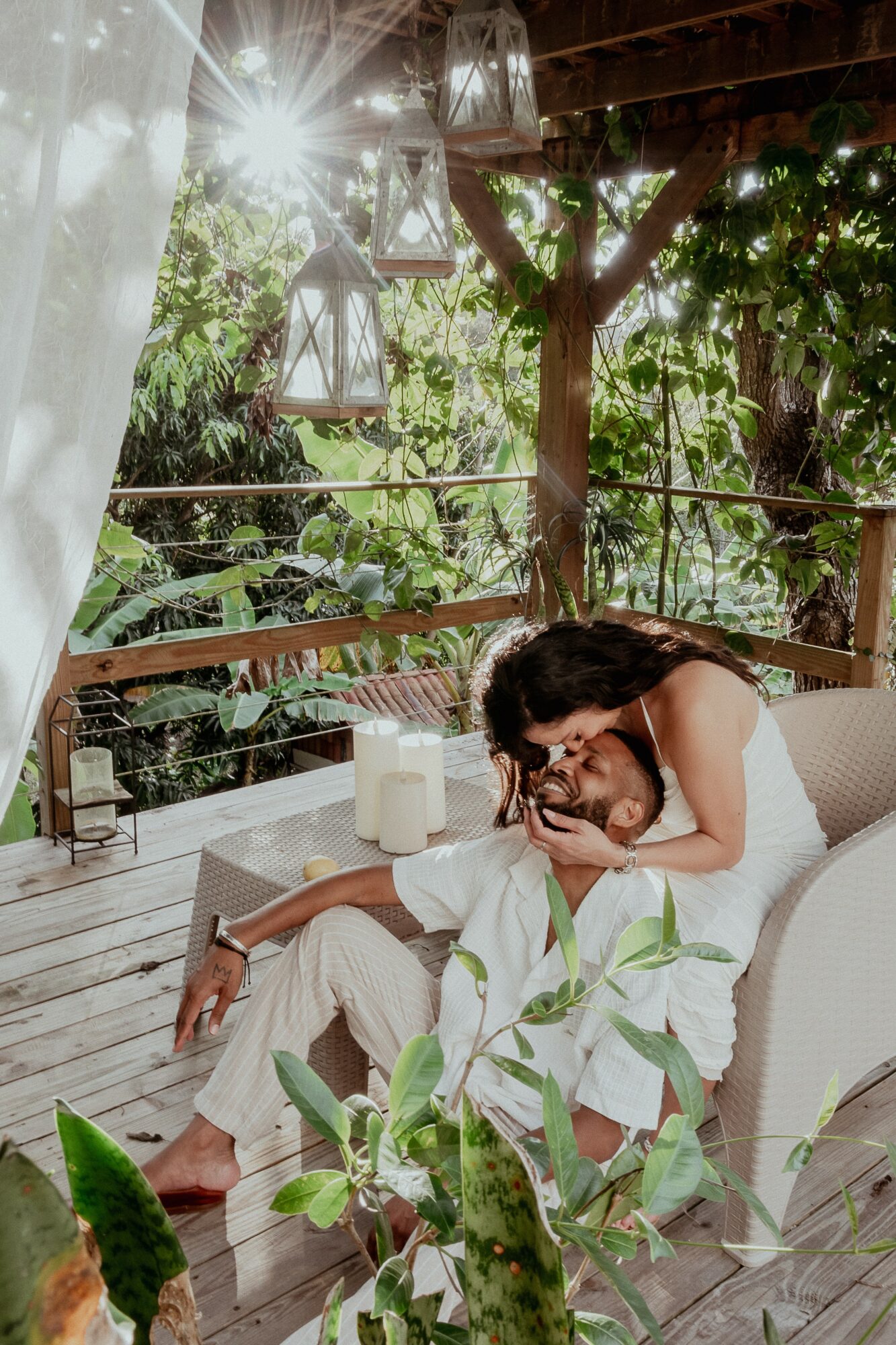 Two people sitting on a wooden deck with greenery and hanging lanterns in the background, one resting head on the other's shoulder.