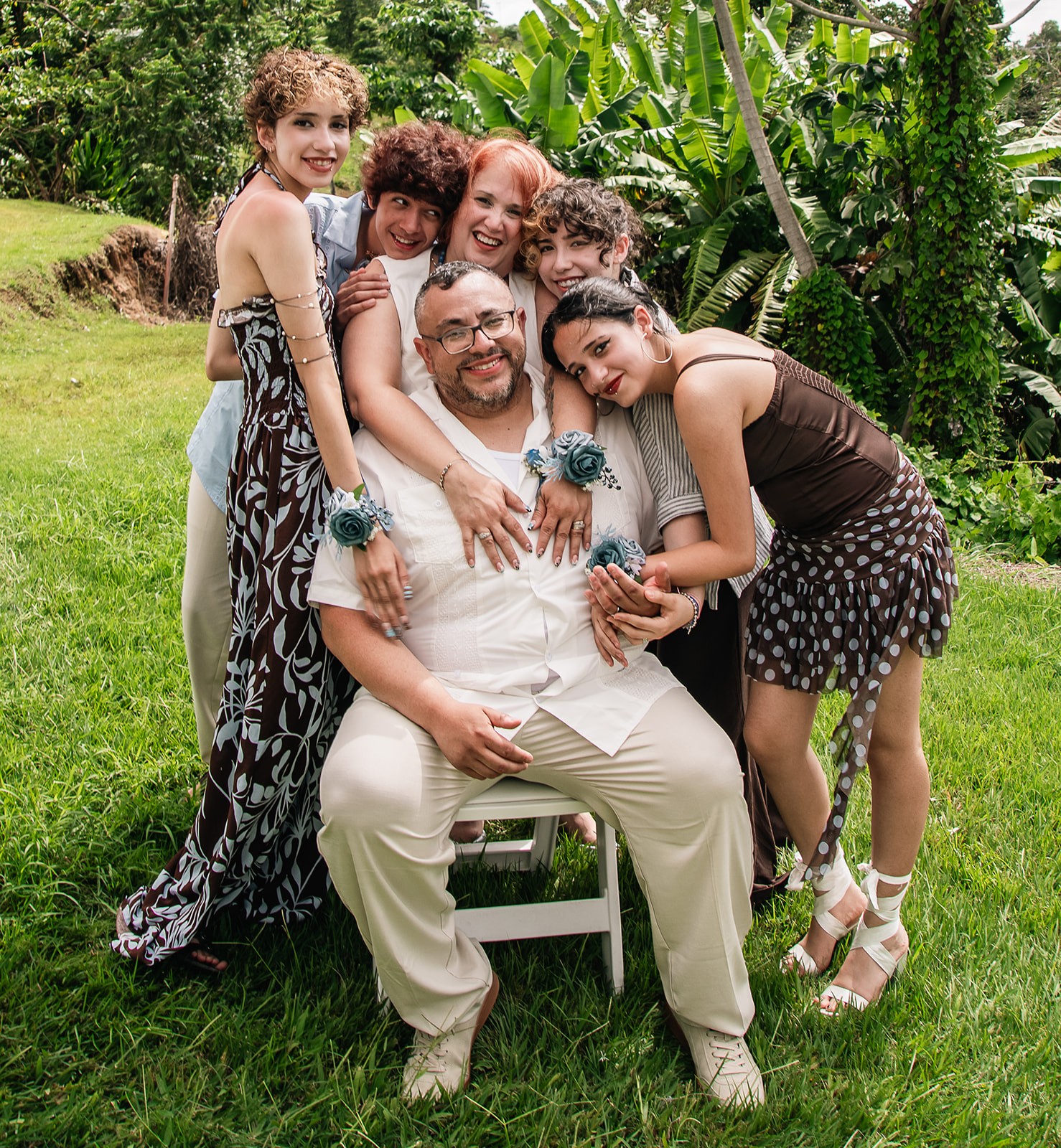 Group of six people outdoors, smiling, hugging, with greenery in background.