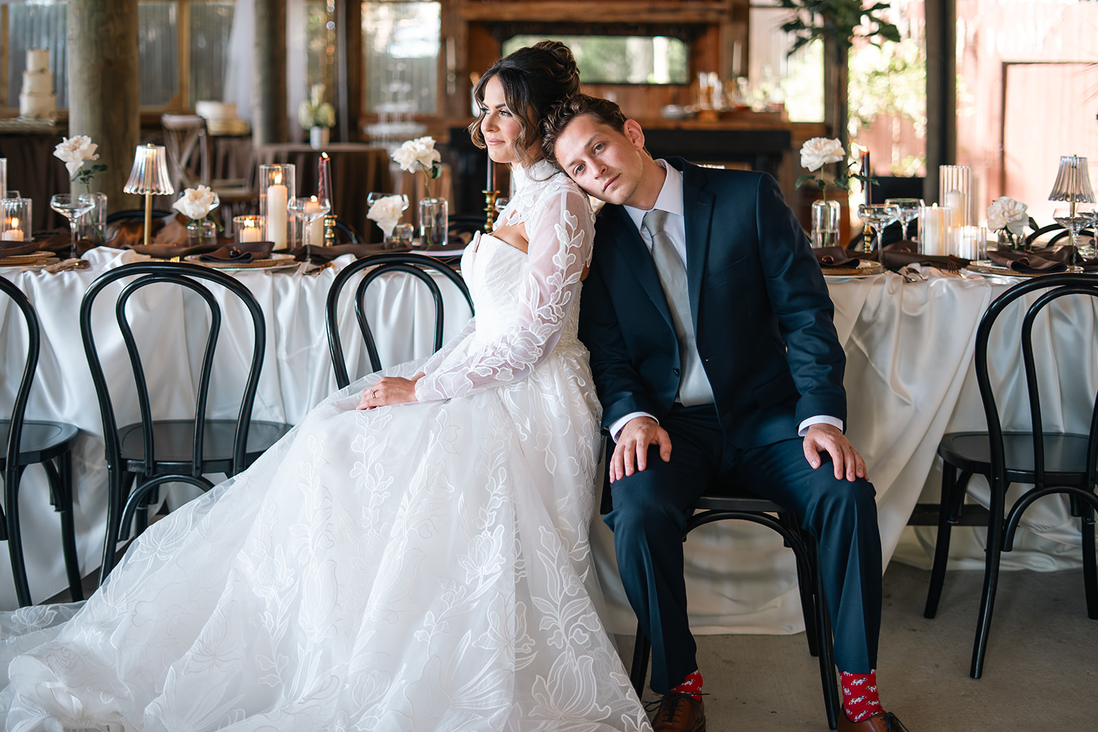 A bride and groom sit together at a decorated wedding reception, with the groom leaning on the bride's shoulder, smiling.
