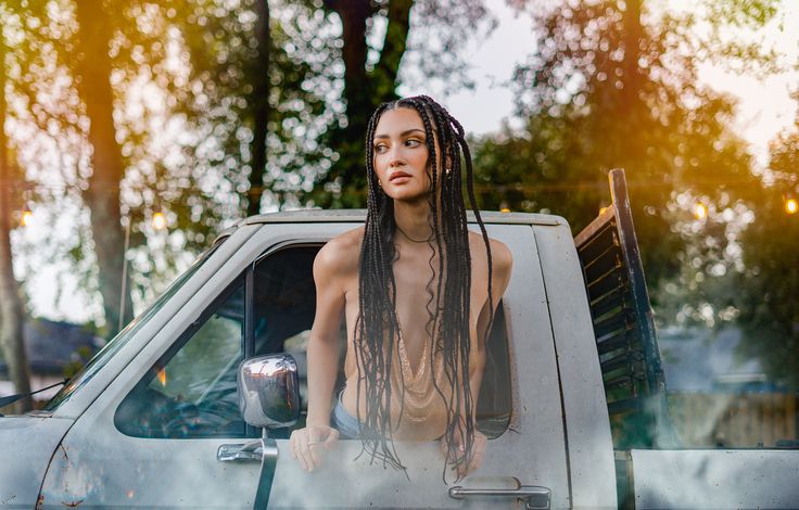 Young woman with long braided hair leaning out of a truck window outdoors, trees and sunlight in background.