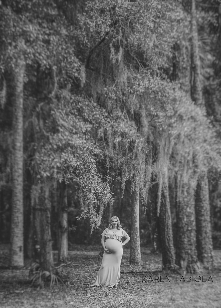 Pregnant woman in a long dress standing outdoors with trees and hanging moss in the background.