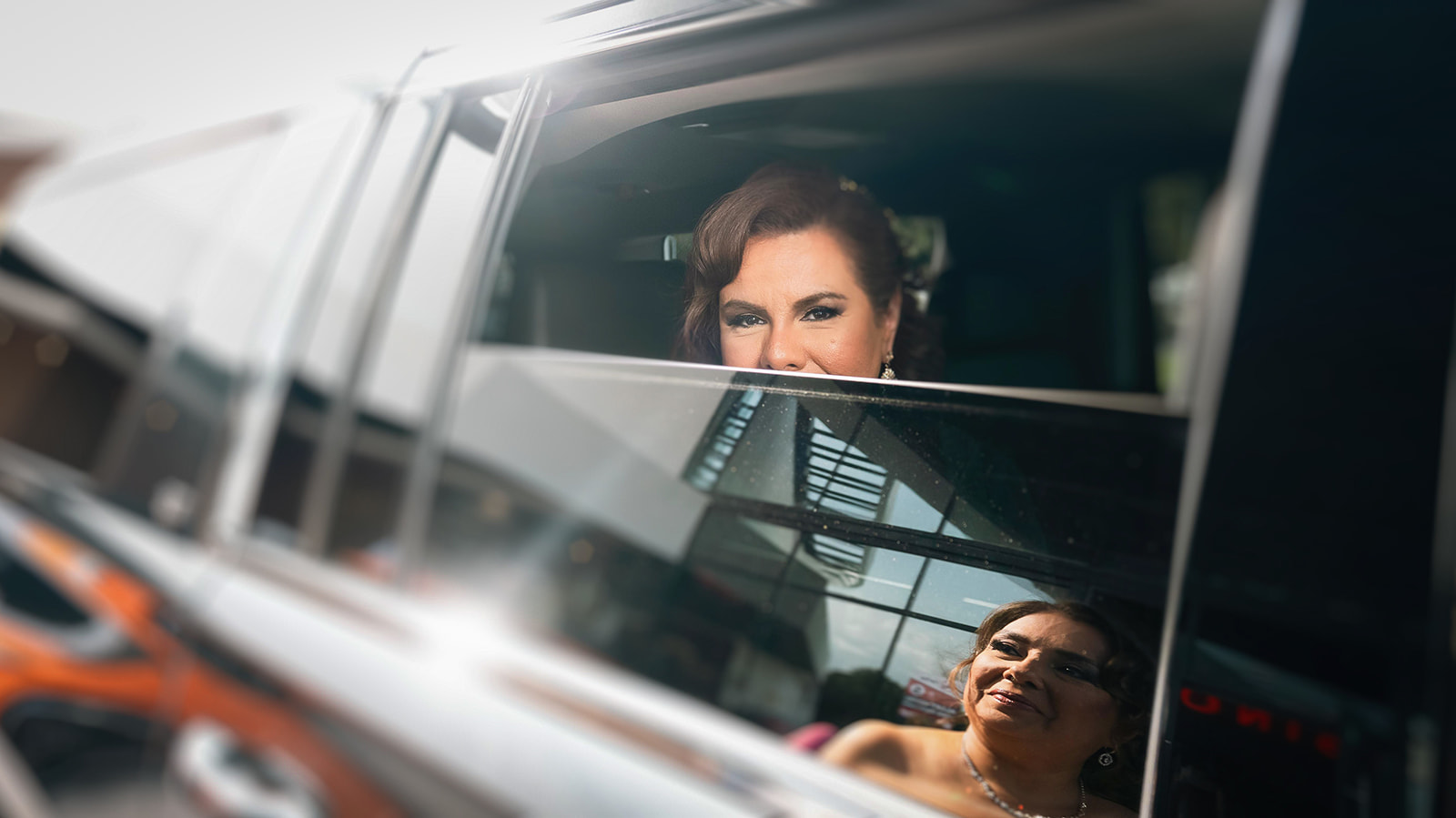 Two women looking out of a car window, one smiling, with reflections of buildings on the glass.