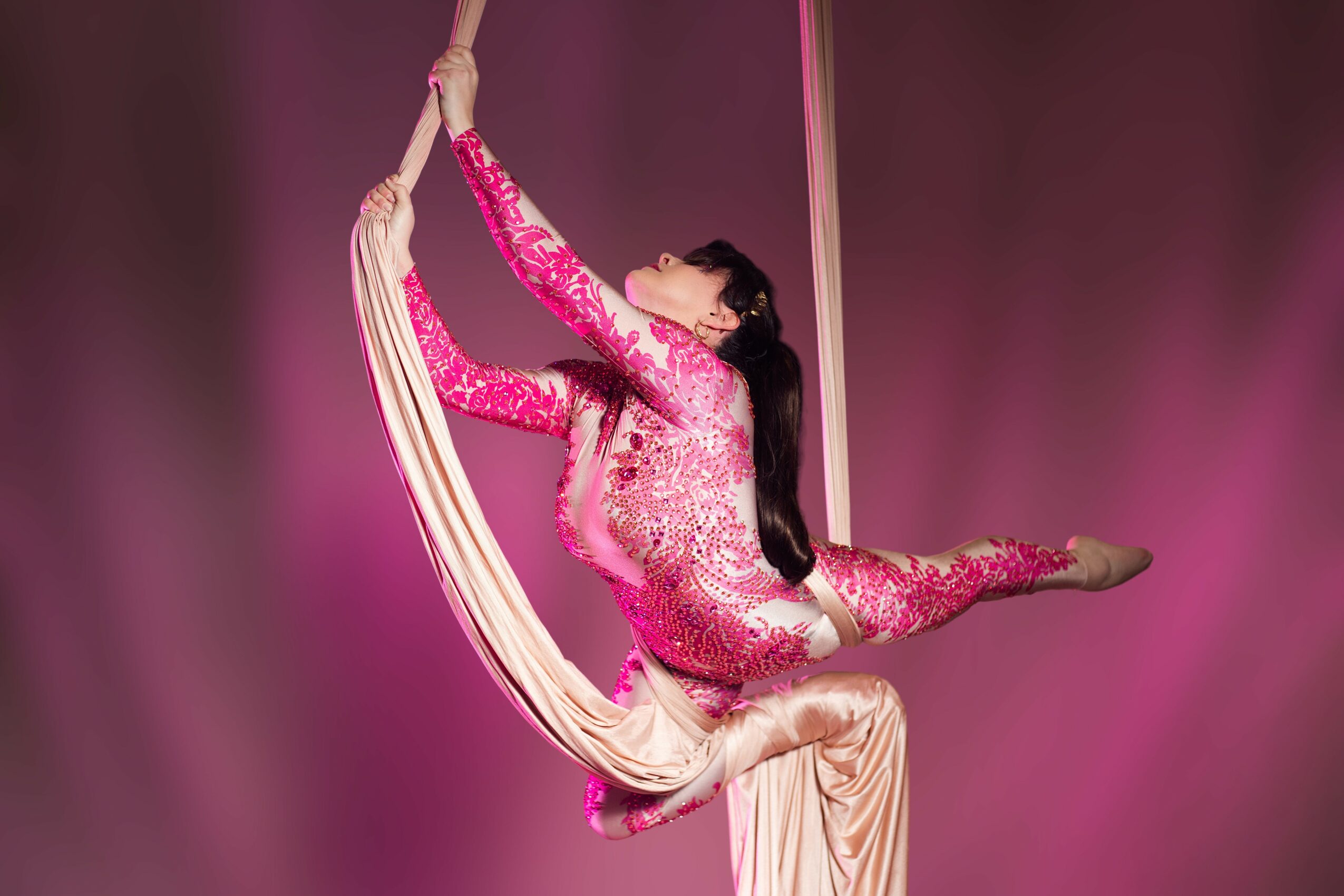 A woman performs an aerial silk routine, holding the fabric with one hand and extending her leg sideways, against a pink background.