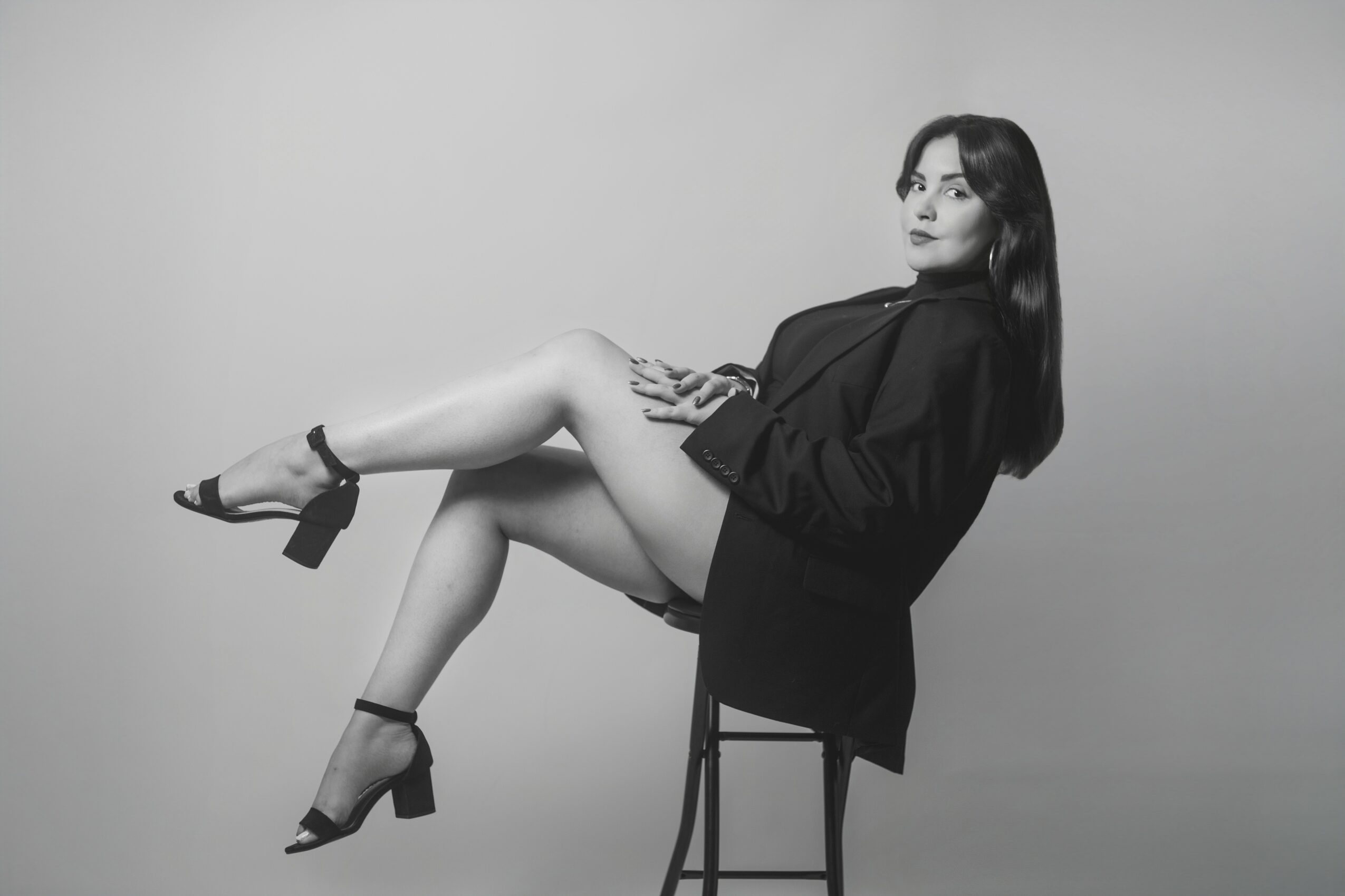 Woman sitting sideways on a chair, legs crossed, wearing a blazer and high heels, looking to the side.