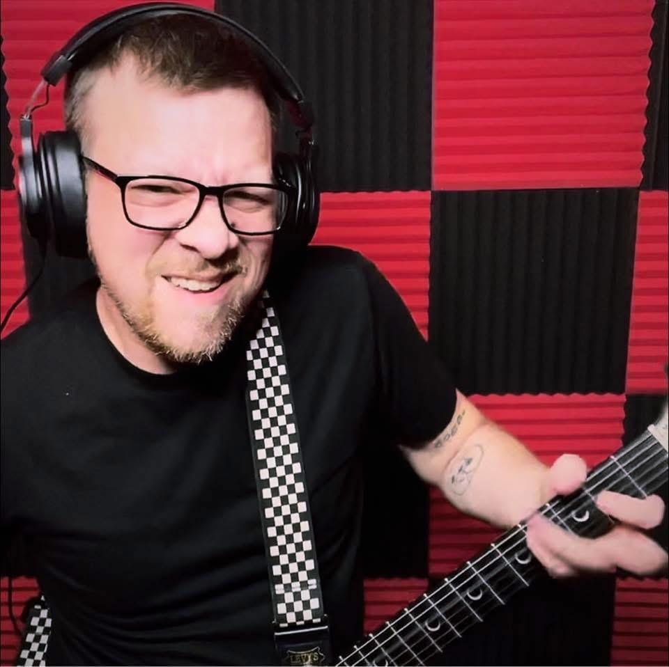 Person with glasses and headphones playing guitar in a soundproof room with red and black foam panels.