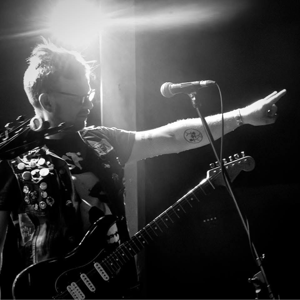Person with glasses and tattoos pointing with one arm while holding a guitar, microphone nearby, in a dark setting.