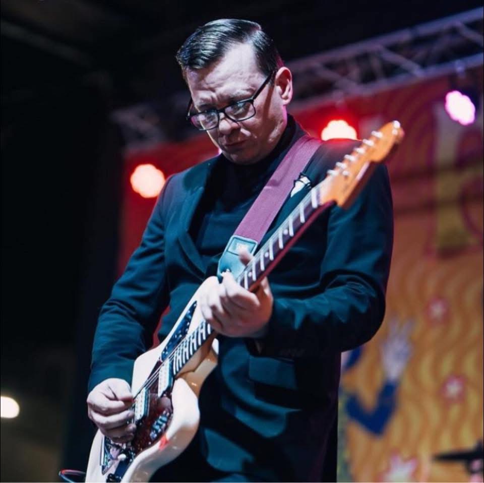 Man playing electric guitar on stage with colorful background, wearing glasses and dark clothing.