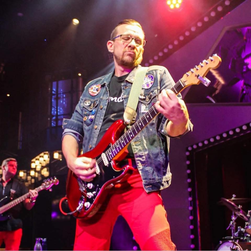 Man playing red electric guitar on stage with colorful lights, wearing a denim jacket with patches and red pants.