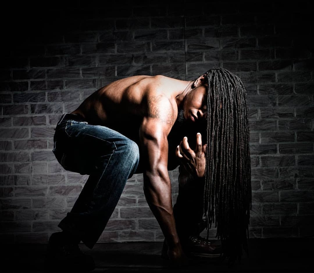 Person with long dreadlocks crouching shirtless against a brick wall, head bowed, hand near face.