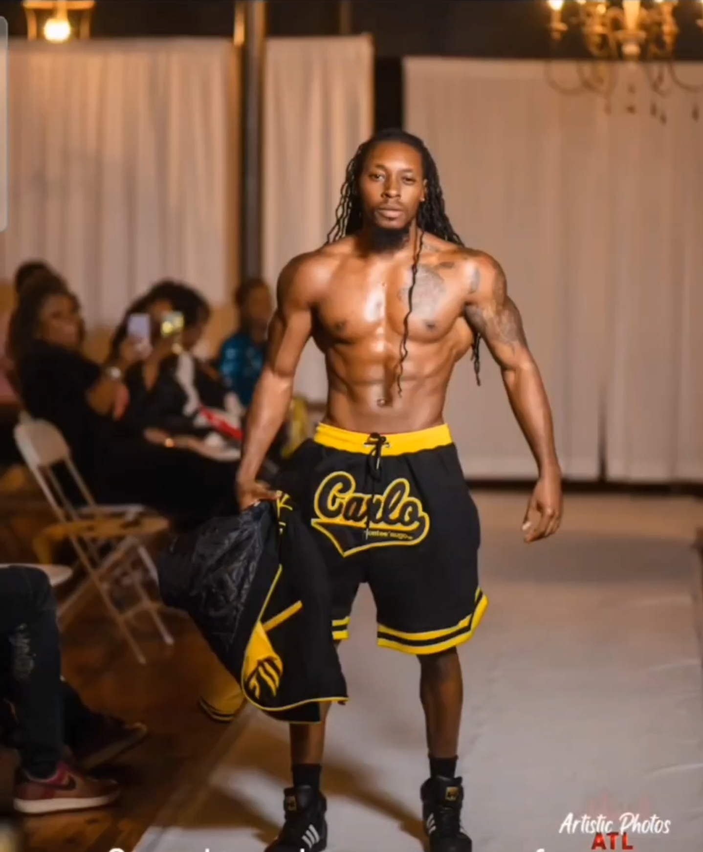 Male model walking on runway with audience in background, wearing black shorts with yellow accents, holding a black jacket, shirtless, muscular, long hair