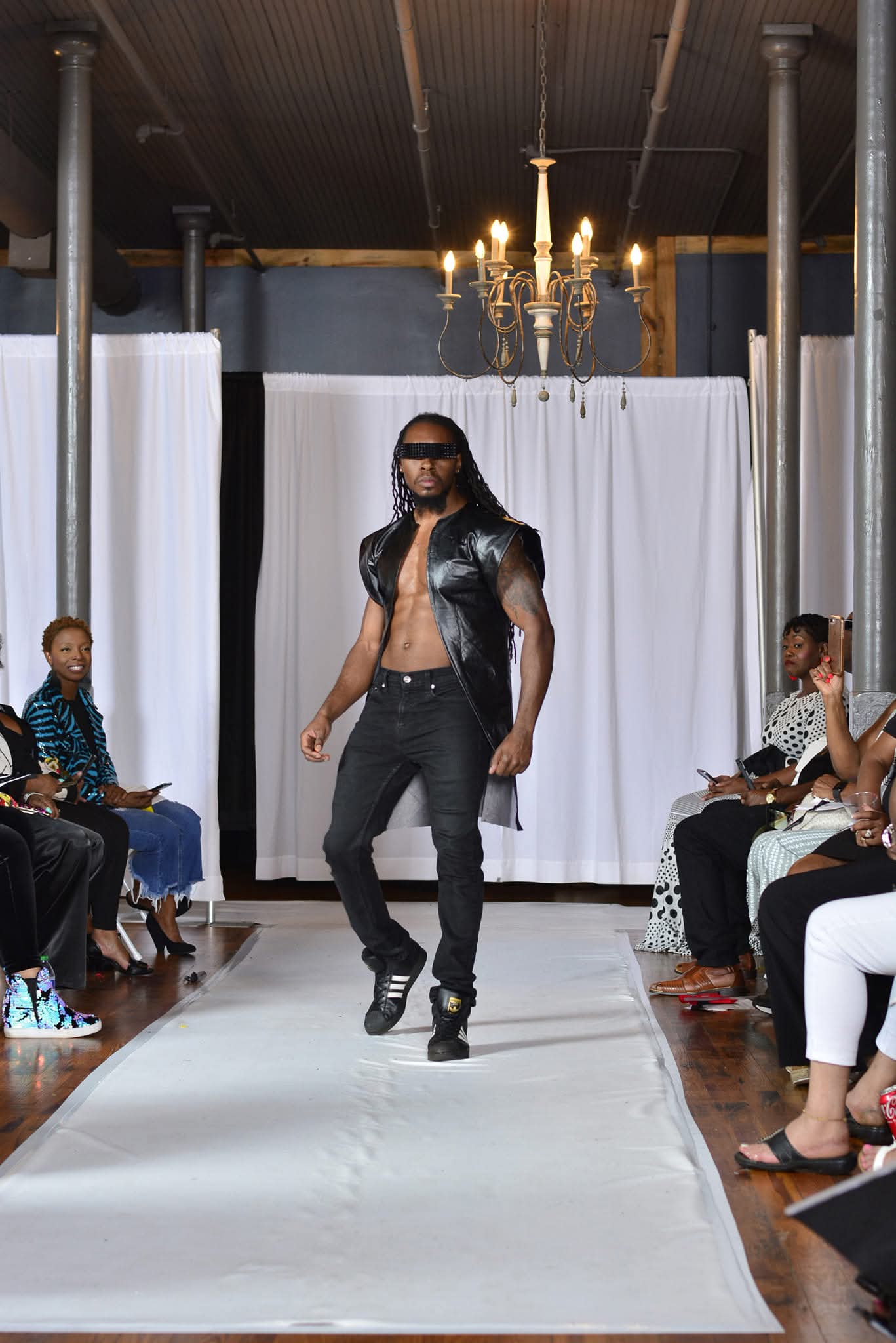 Male model walking on runway with audience on both sides, white backdrop, chandelier overhead, and industrial ceiling.