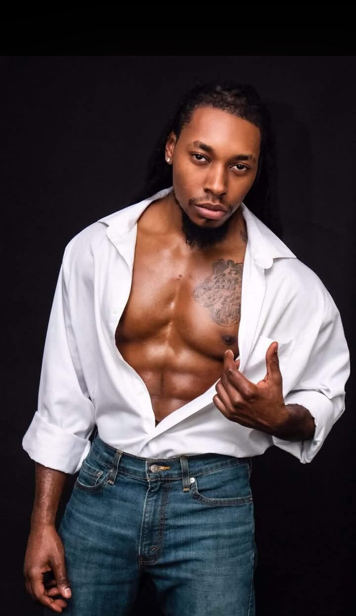 Man with open white shirt, jeans, and tattoos on chest, posing against black background, making a hand gesture.