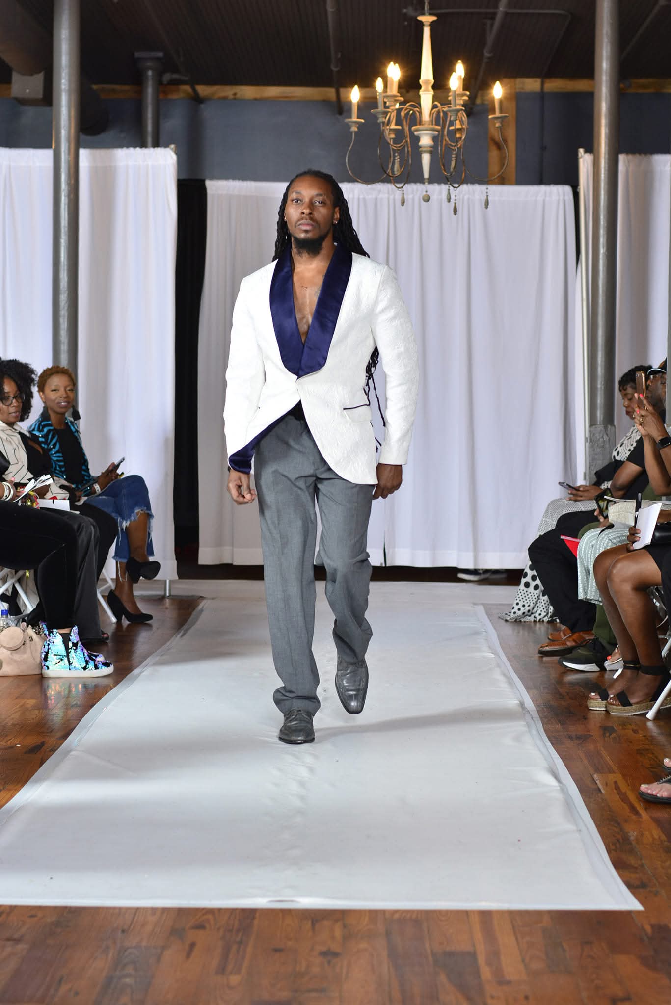Male model walking down runway with audience on both sides, white backdrop, chandelier overhead, wearing a white blazer and gray pants.