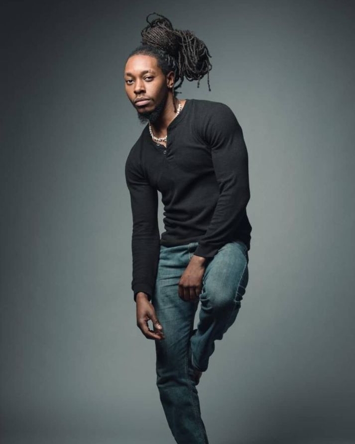 Man with dreadlocks wearing a black long-sleeve shirt and jeans, standing against a plain gray background.