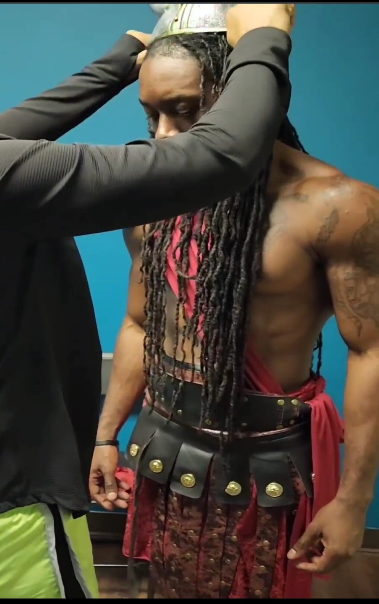 Person with long dreadlocks and muscular build, wearing a red cloth around waist, standing against a blue background.