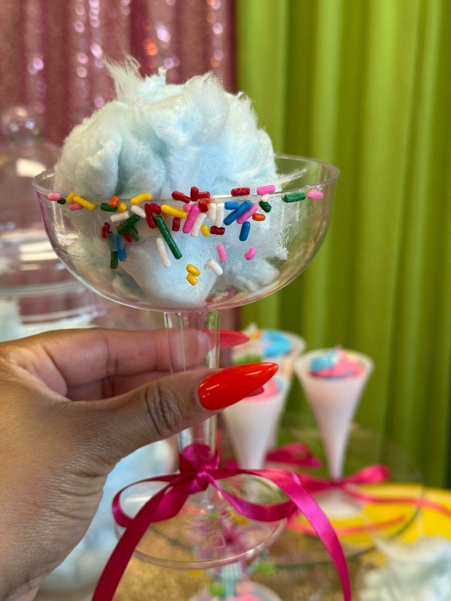 Person holding a cotton candy with colorful sprinkles in a clear plastic cup, with party hats and decorations in background.