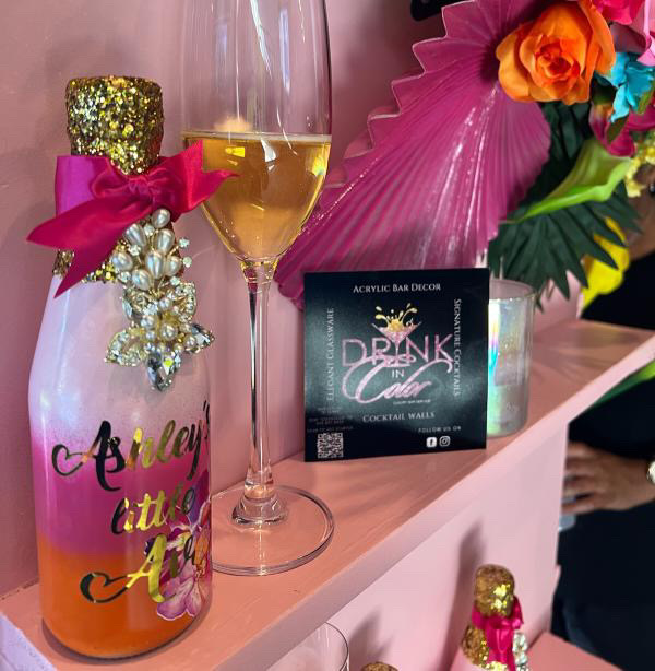 Pink bottle with a pink bow and gold glitter, a glass of white wine, a colorful flower arrangement, and a black sign on a pink surface.