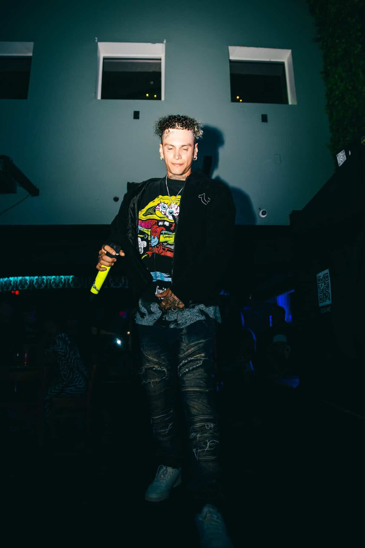 Young man holding a microphone in a dark indoor setting with a modern building background.