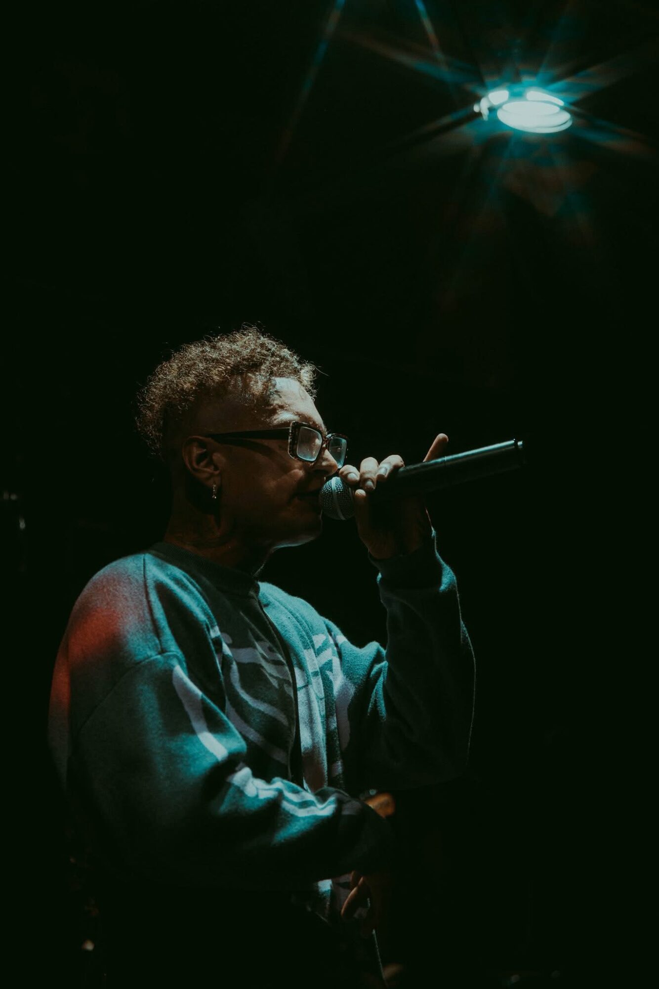 Person with curly hair and glasses singing into a microphone under a spotlight in a dark setting.