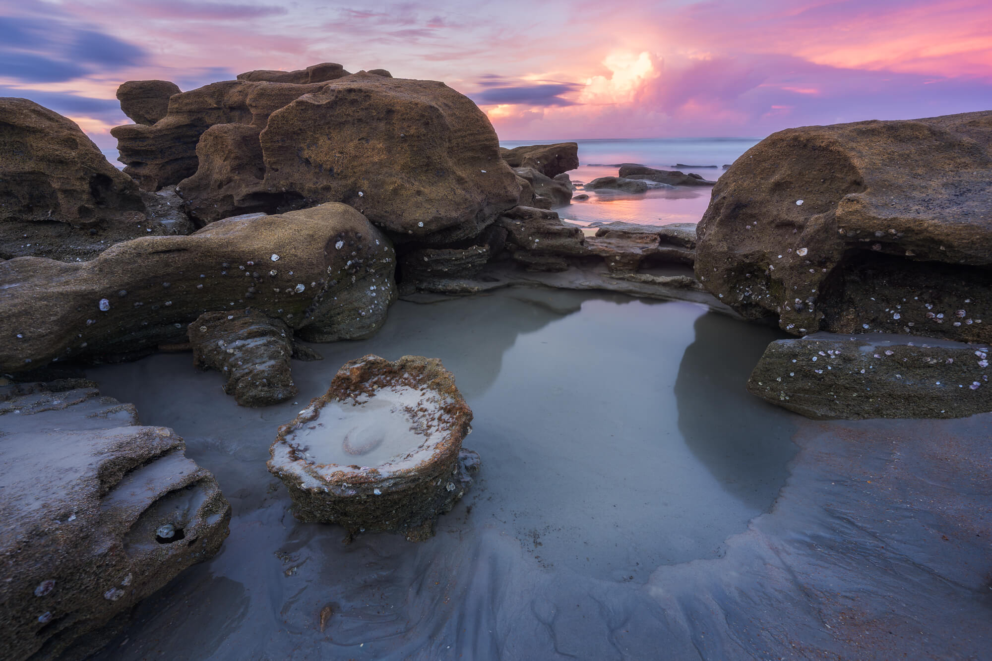 Rock formations on a beach with water and a colorful sunset sky in the background.