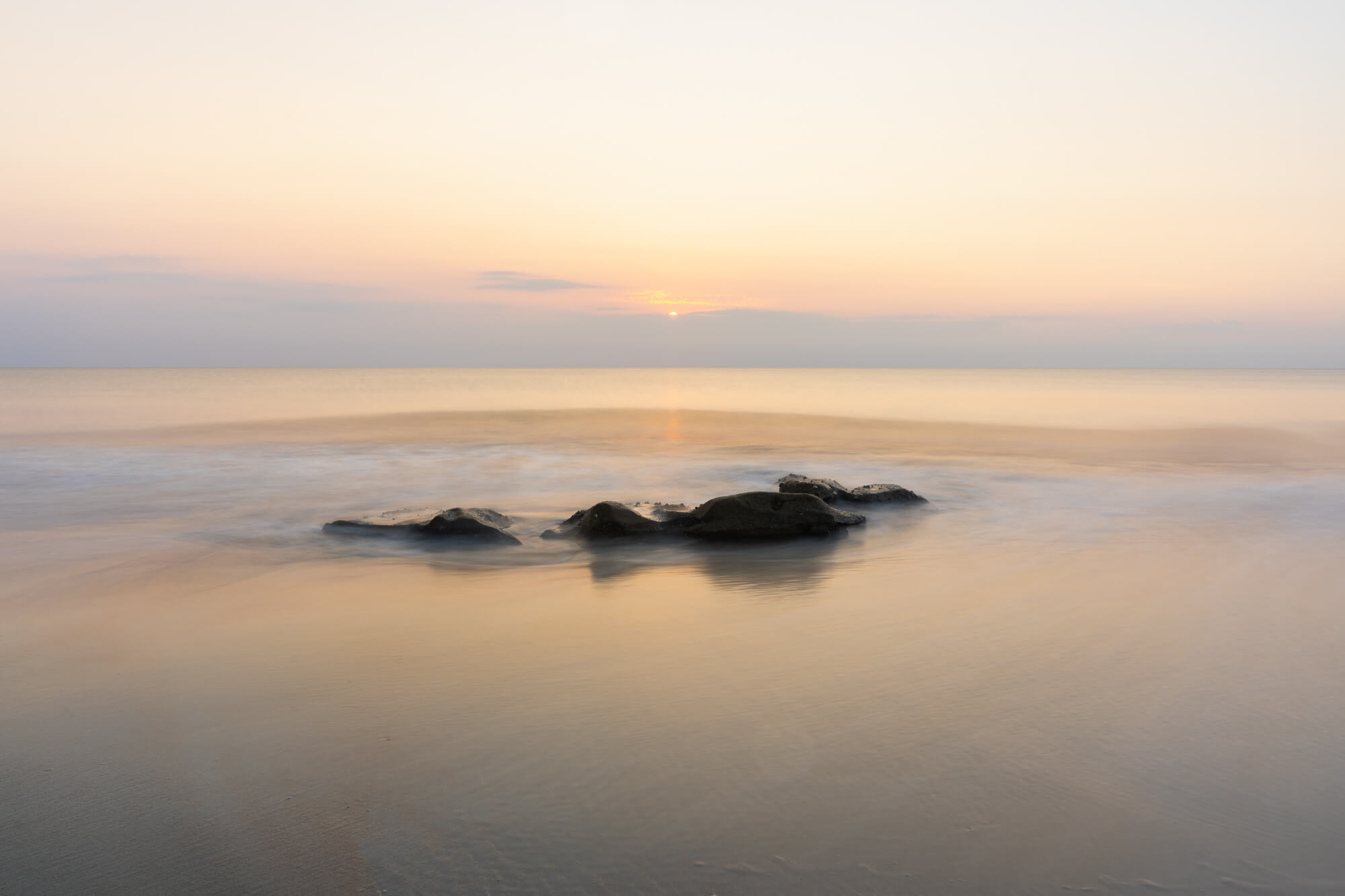 Calm ocean with rocks in water during sunset, soft pastel sky, minimal waves, tranquil scene