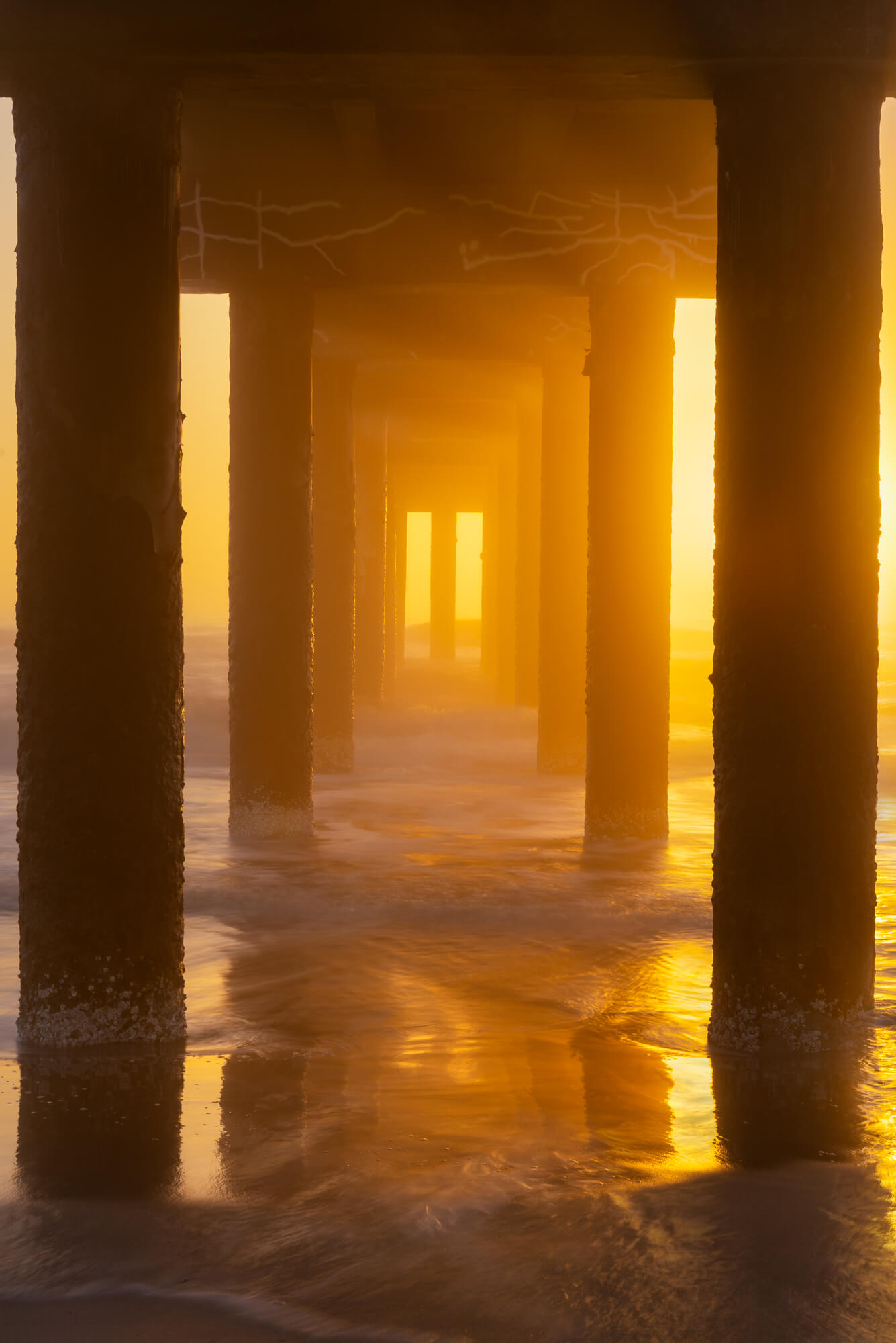 View underneath a pier with columns extending into the ocean, illuminated by golden sunlight, reflecting on wet sand.
